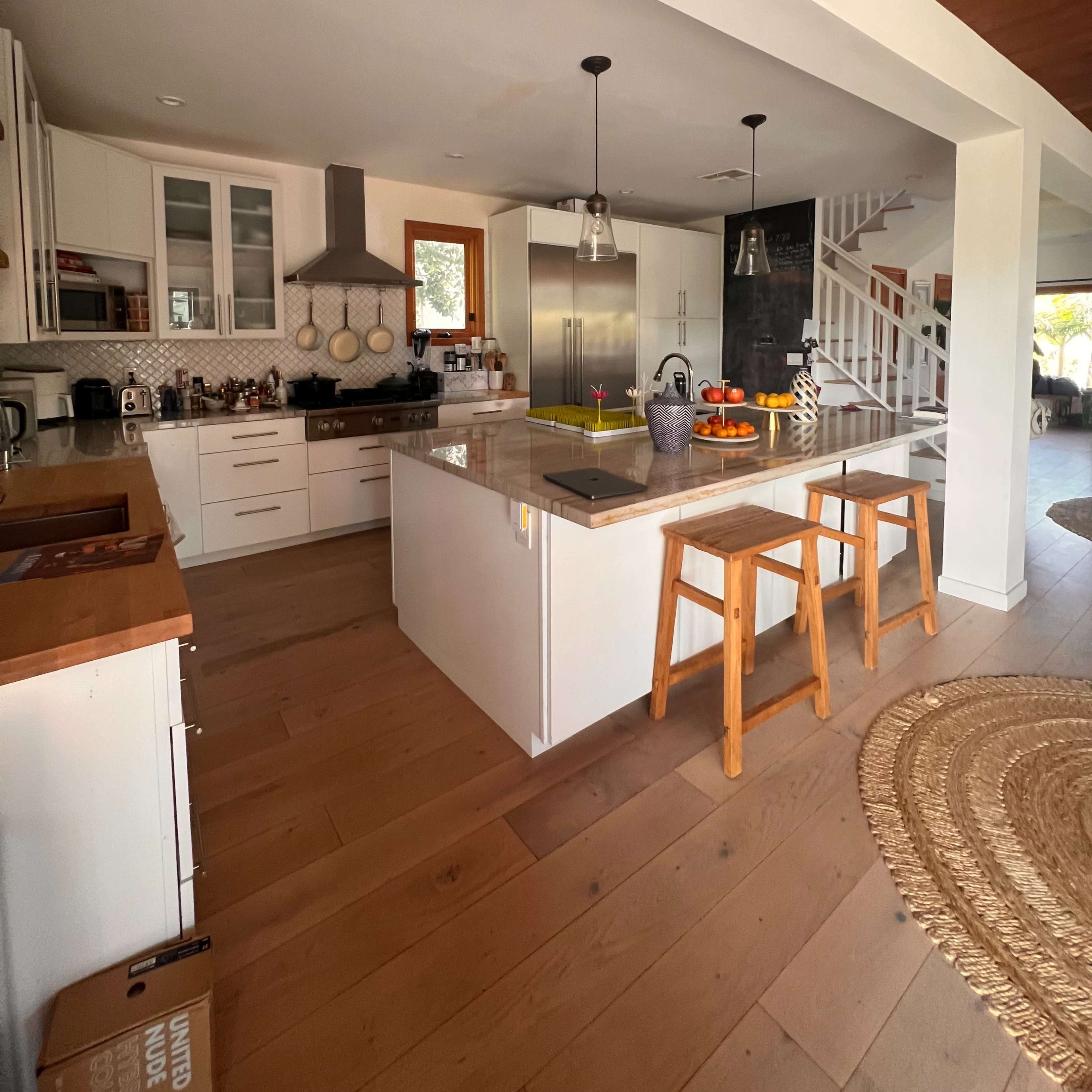 A modern kitchen features a central island with wooden stools, sleek cabinetry, and a mix of natural light and shadows from overhead lighting.