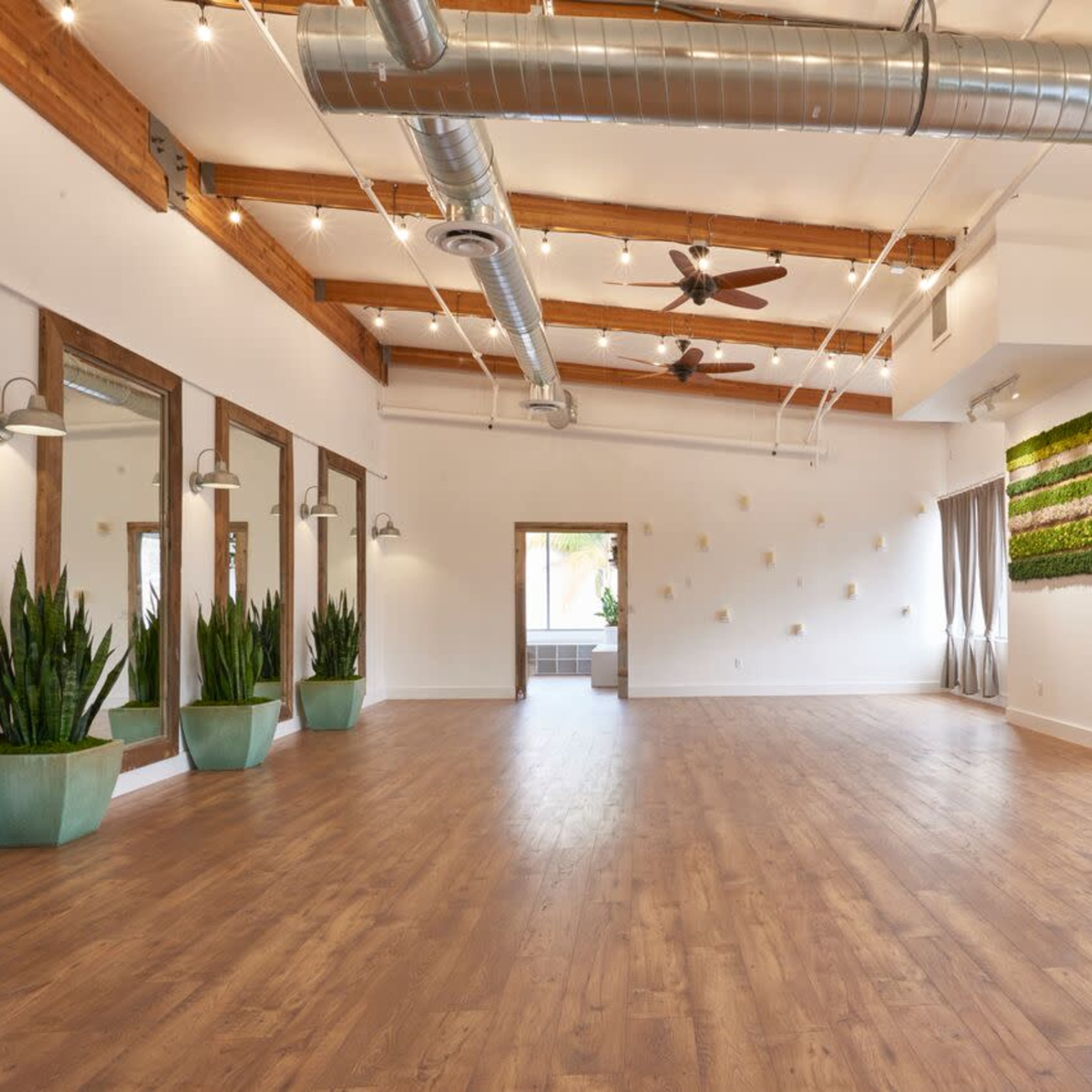 An empty interior space features large mirrors, potted plants, and a wooden floor with exposed beams and modern lighting.