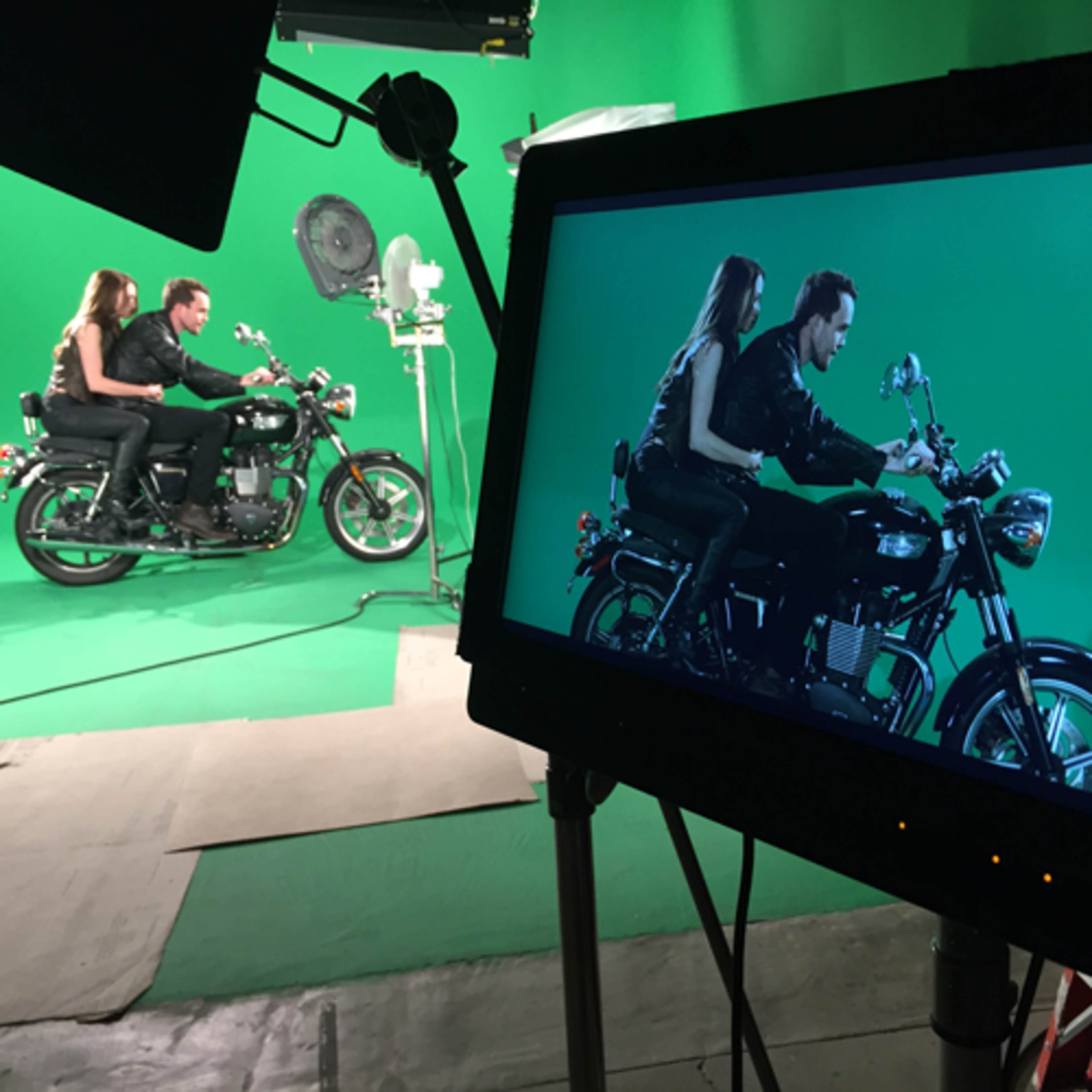 A couple sits on a motorcycle in front of a green screen while being filmed from a monitor.