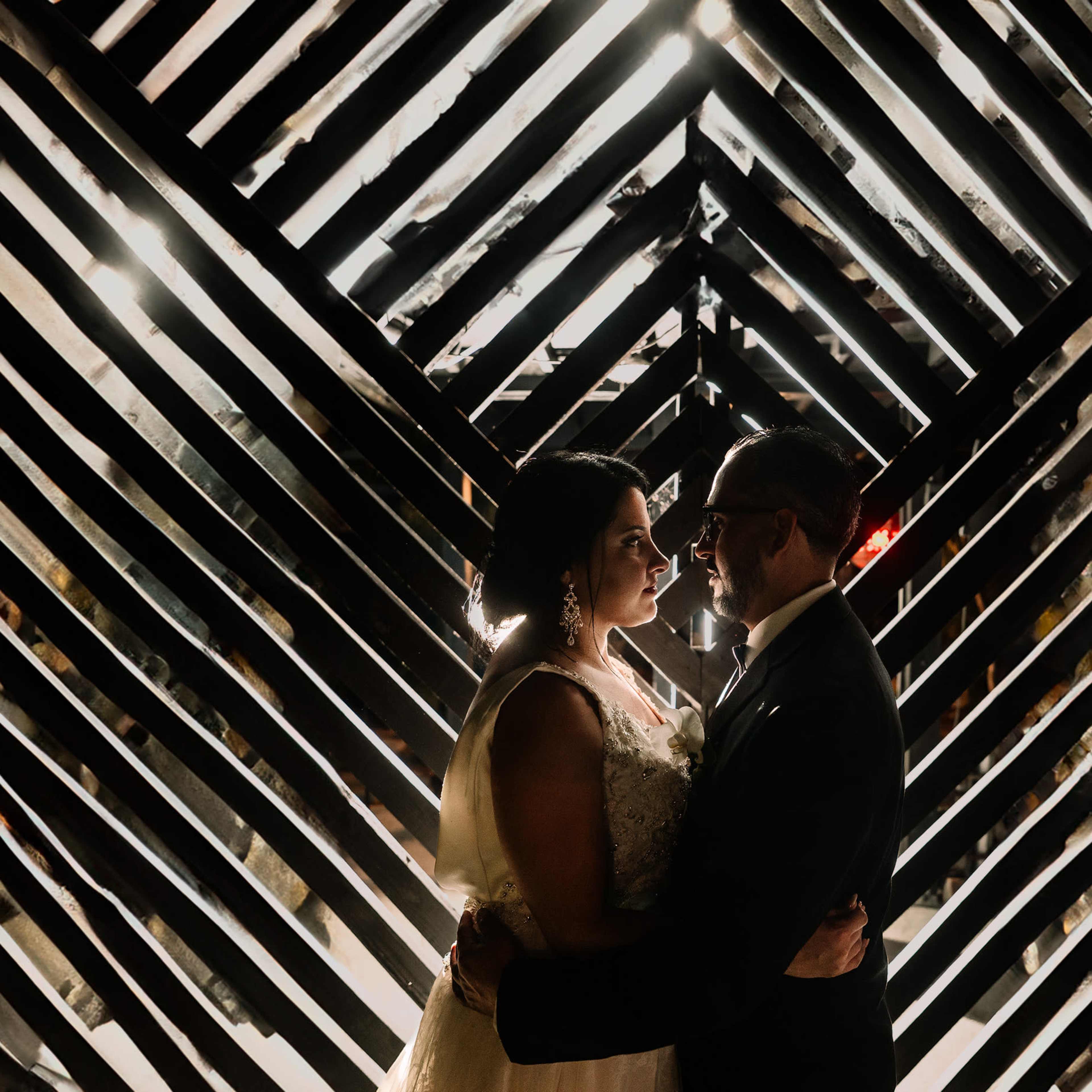 A couple stands embracing in front of a dramatic backdrop of intersecting black wooden slats.