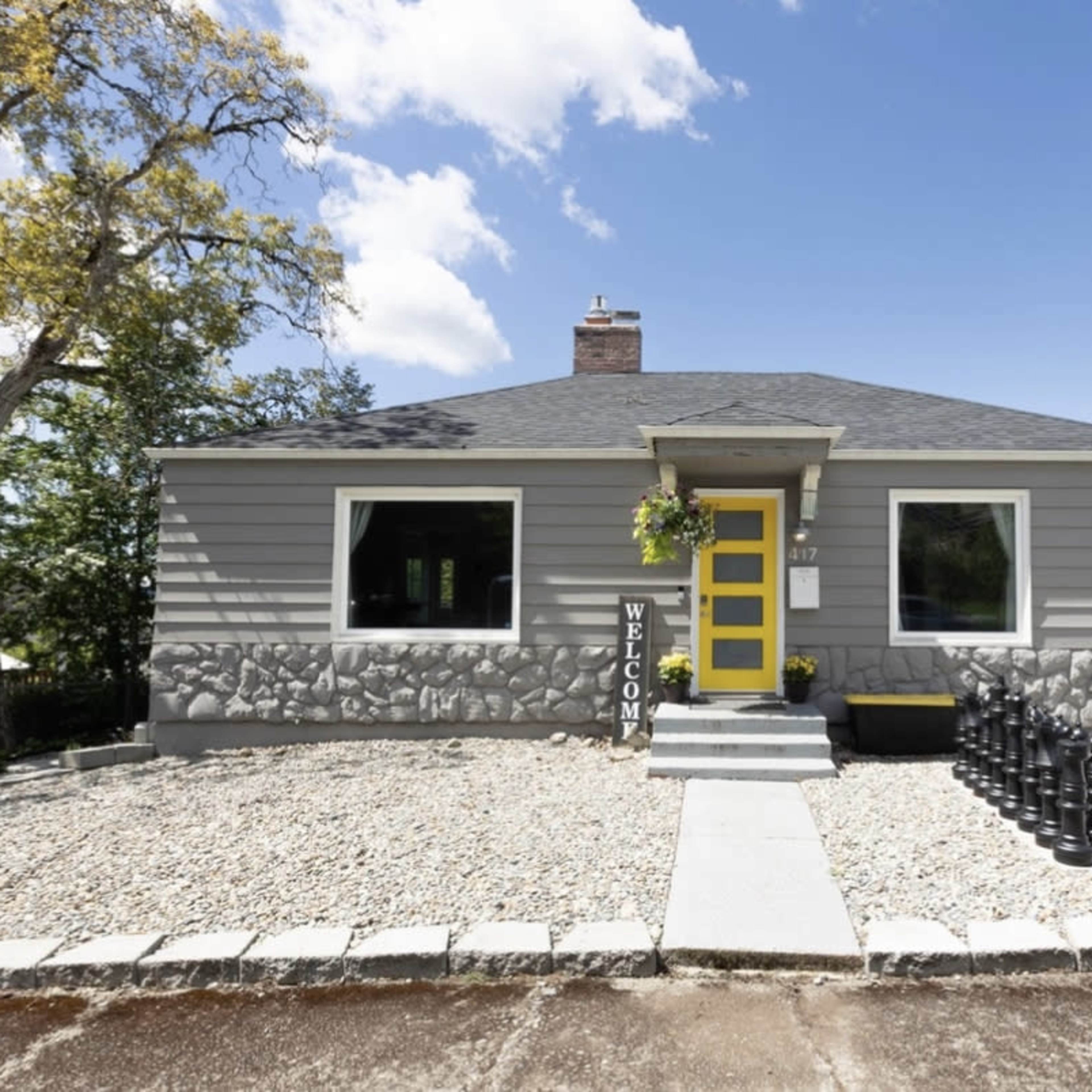 A single-story house with gray siding and a yellow front door is flanked by a large decorative chess set on one side and a welcome sign on the other.