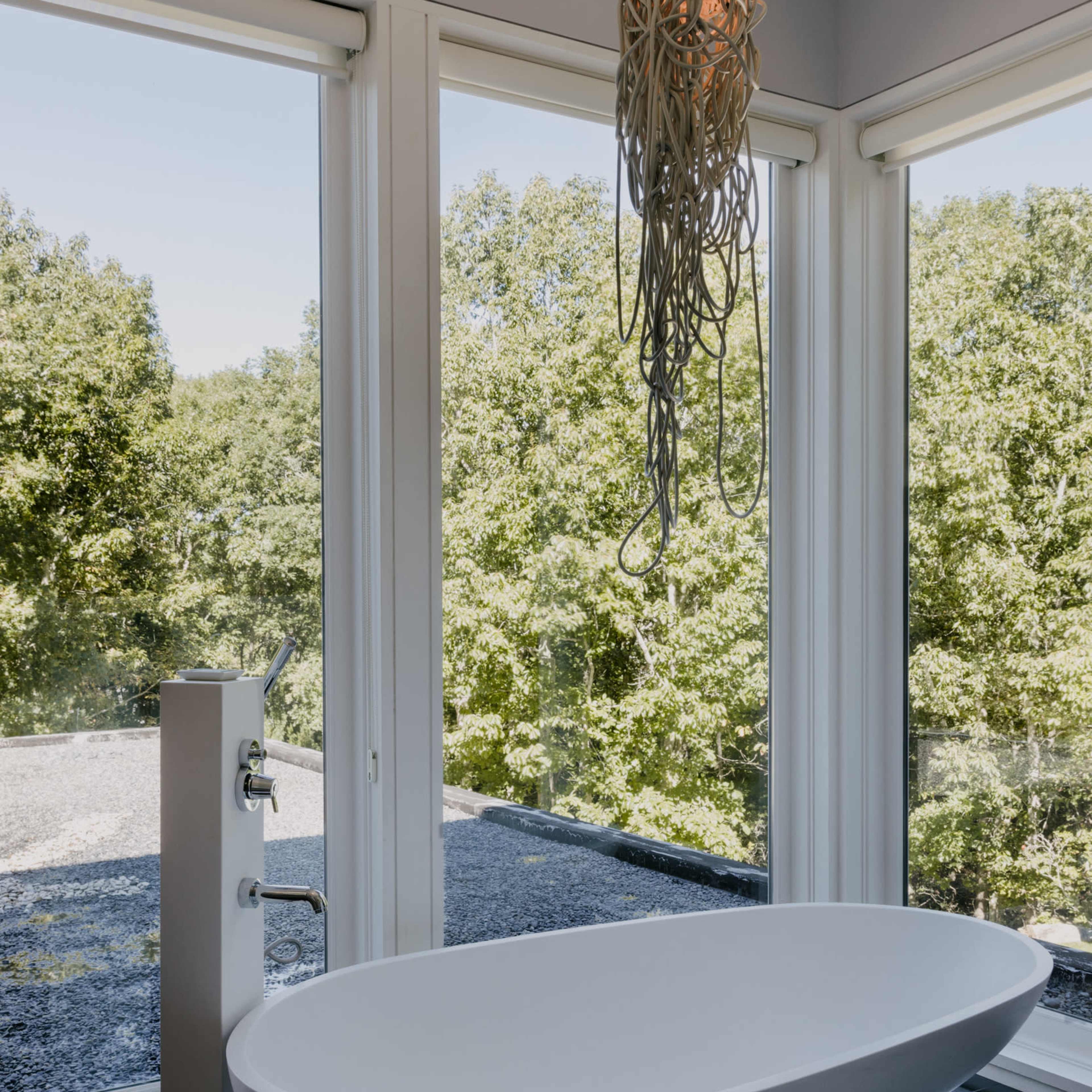 A modern freestanding bathtub is positioned beside large windows, offering a view of a green landscape and featuring a unique hanging light fixture above.