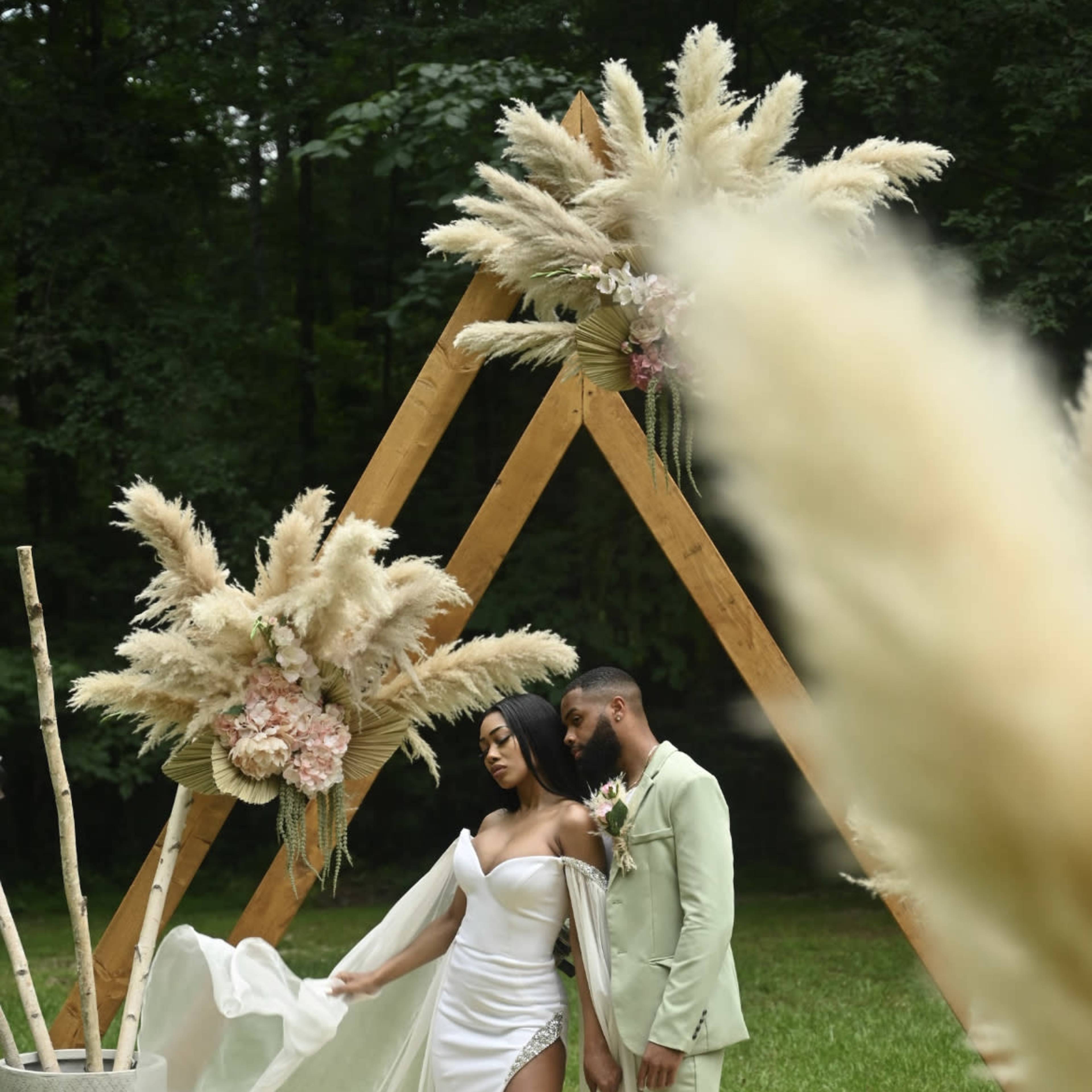 A couple poses together on a round mat beneath an arch adorned with pampas grass in a wooded setting.