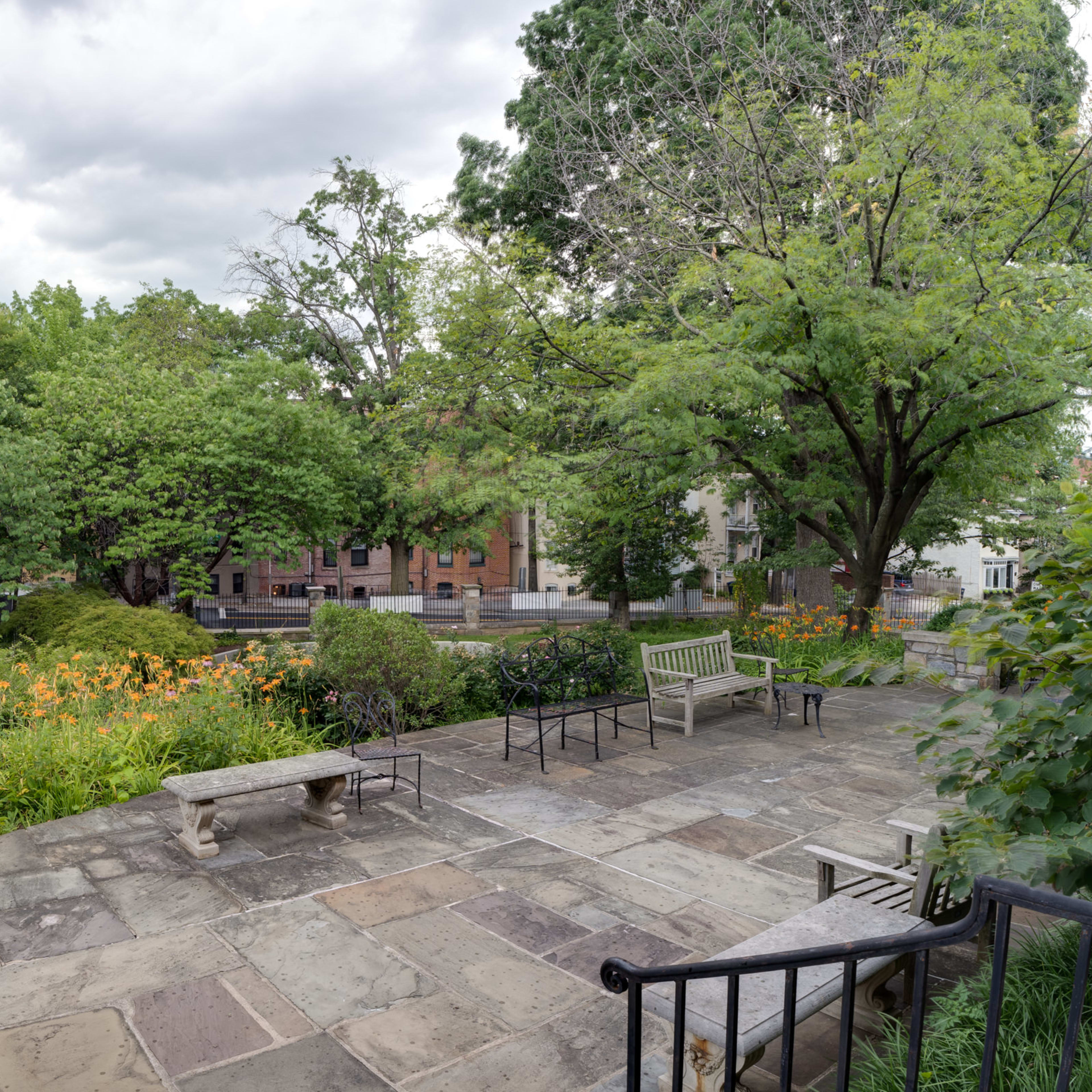 A stone patio with benches overlooks a garden filled with green trees and colorful flowers.