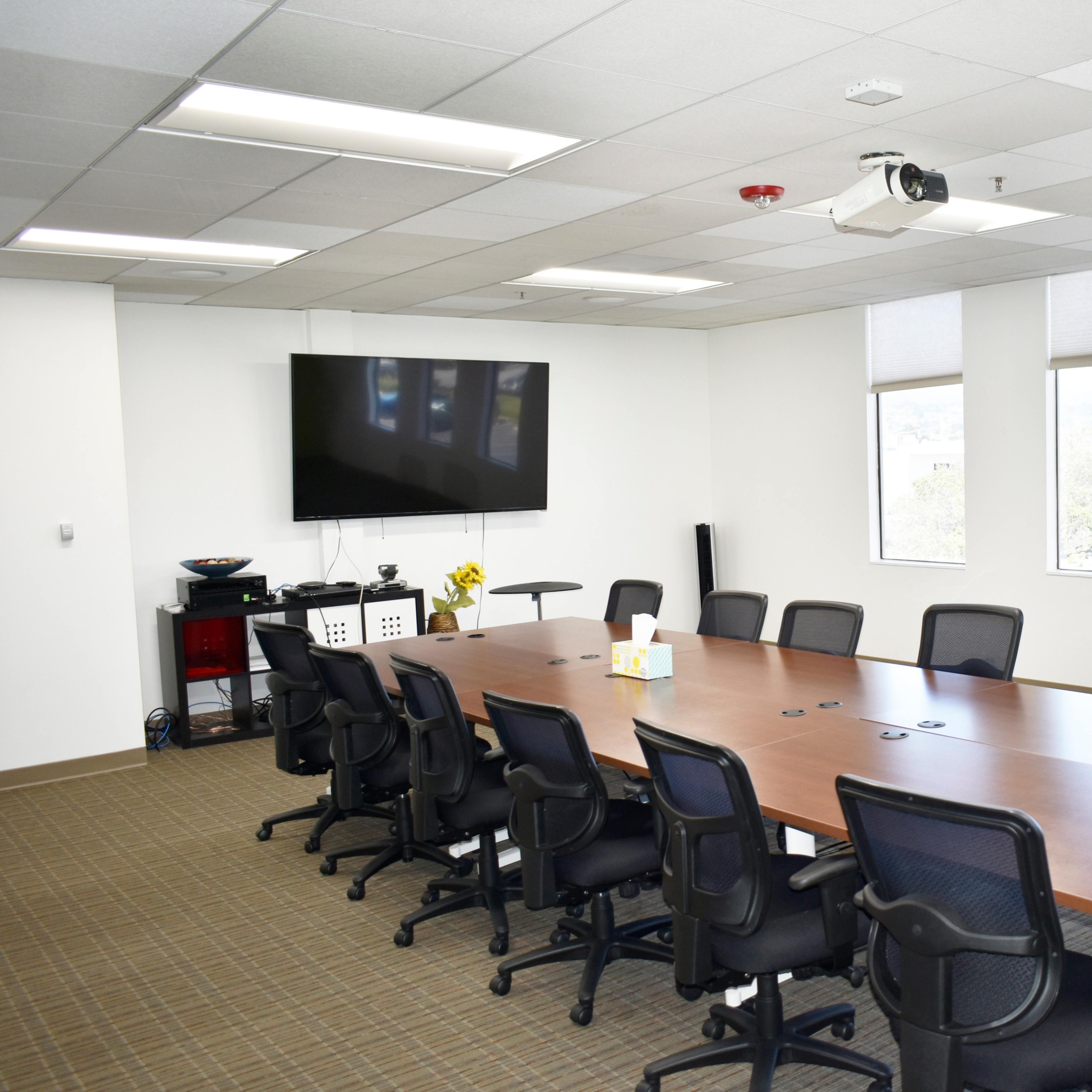 A modern conference room featuring a long wooden table surrounded by black chairs, a large TV screen on the wall, and windows letting in natural light.