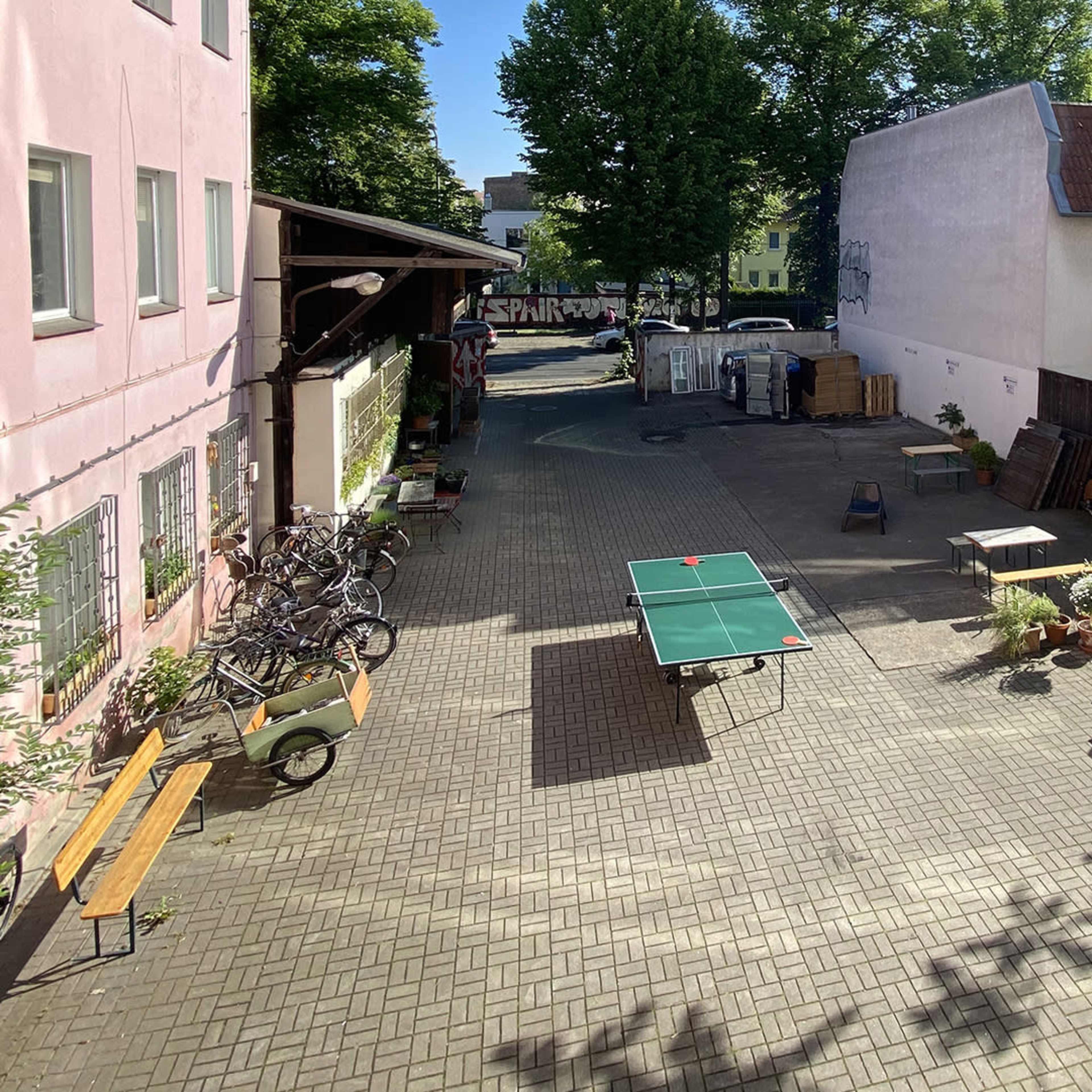 A sunny courtyard with bicycles parked, a ping pong table, and a person sitting at a table surrounded by potted plants.