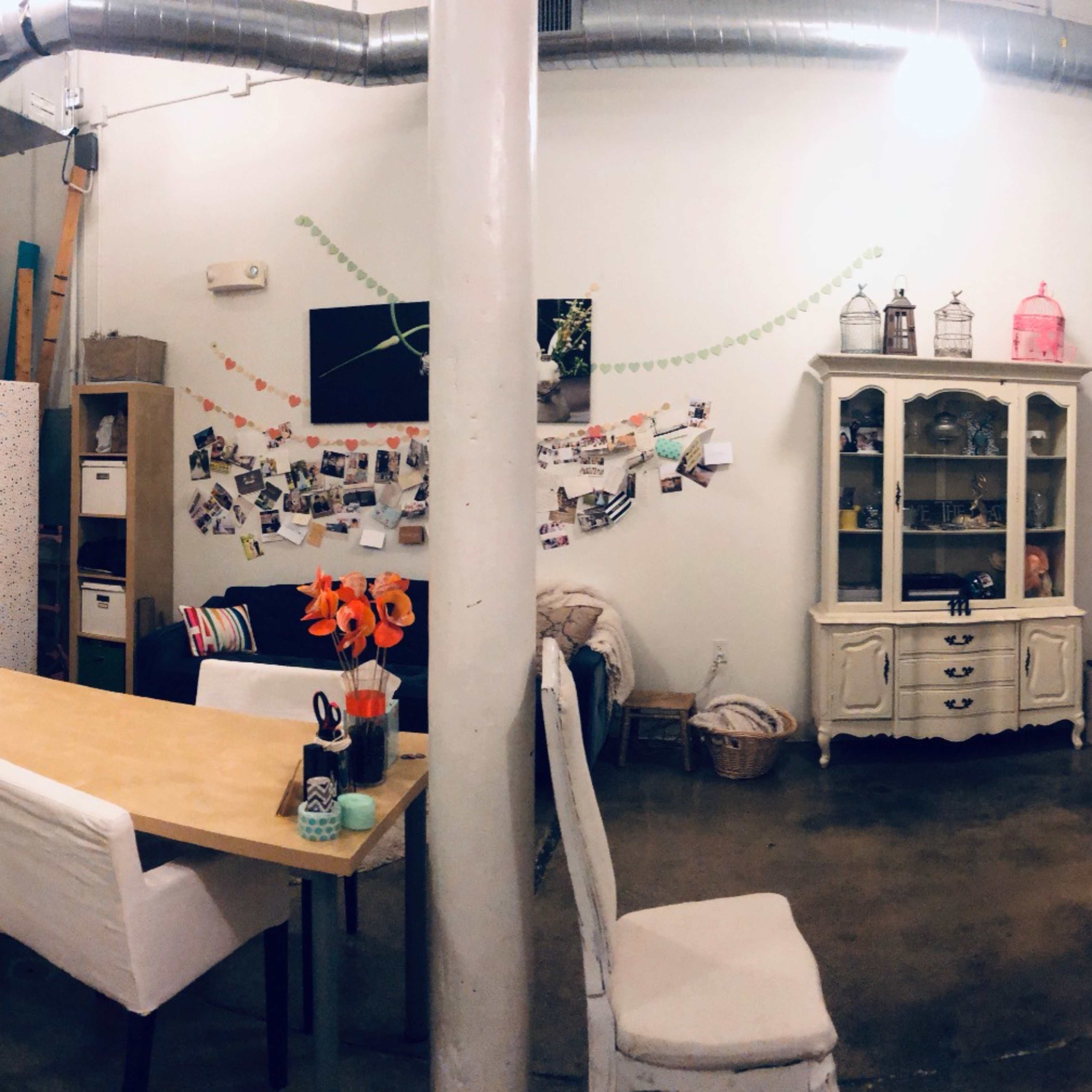 A spacious room with a mix of furniture, including a table and chairs, shelves, and decorative curtains, alongside a large window letting in natural light.
