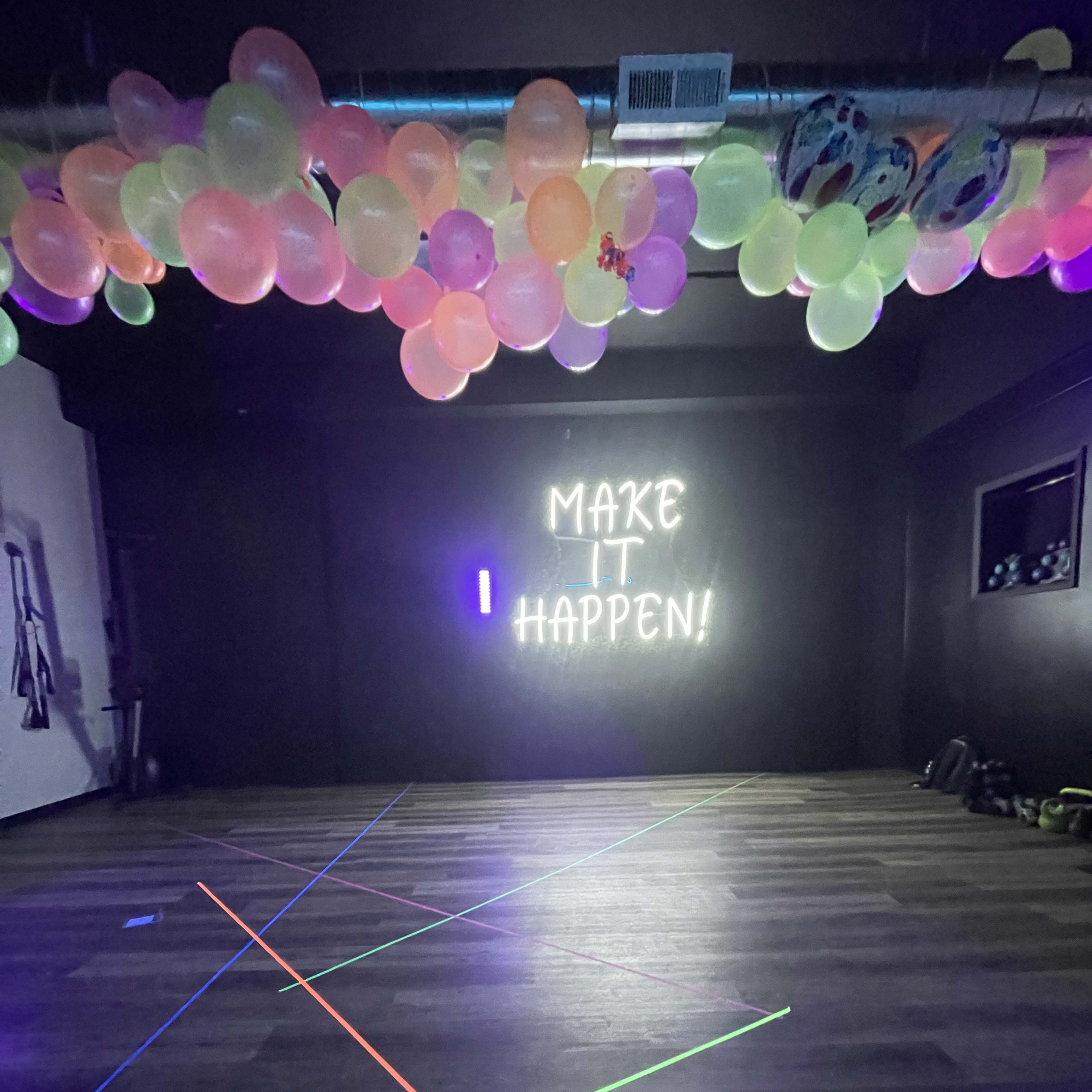 A dimly lit room with colorful balloons hanging from the ceiling and a neon sign on the wall that reads "MAKE IT HAPPEN!" with multicolored lines on the floor.