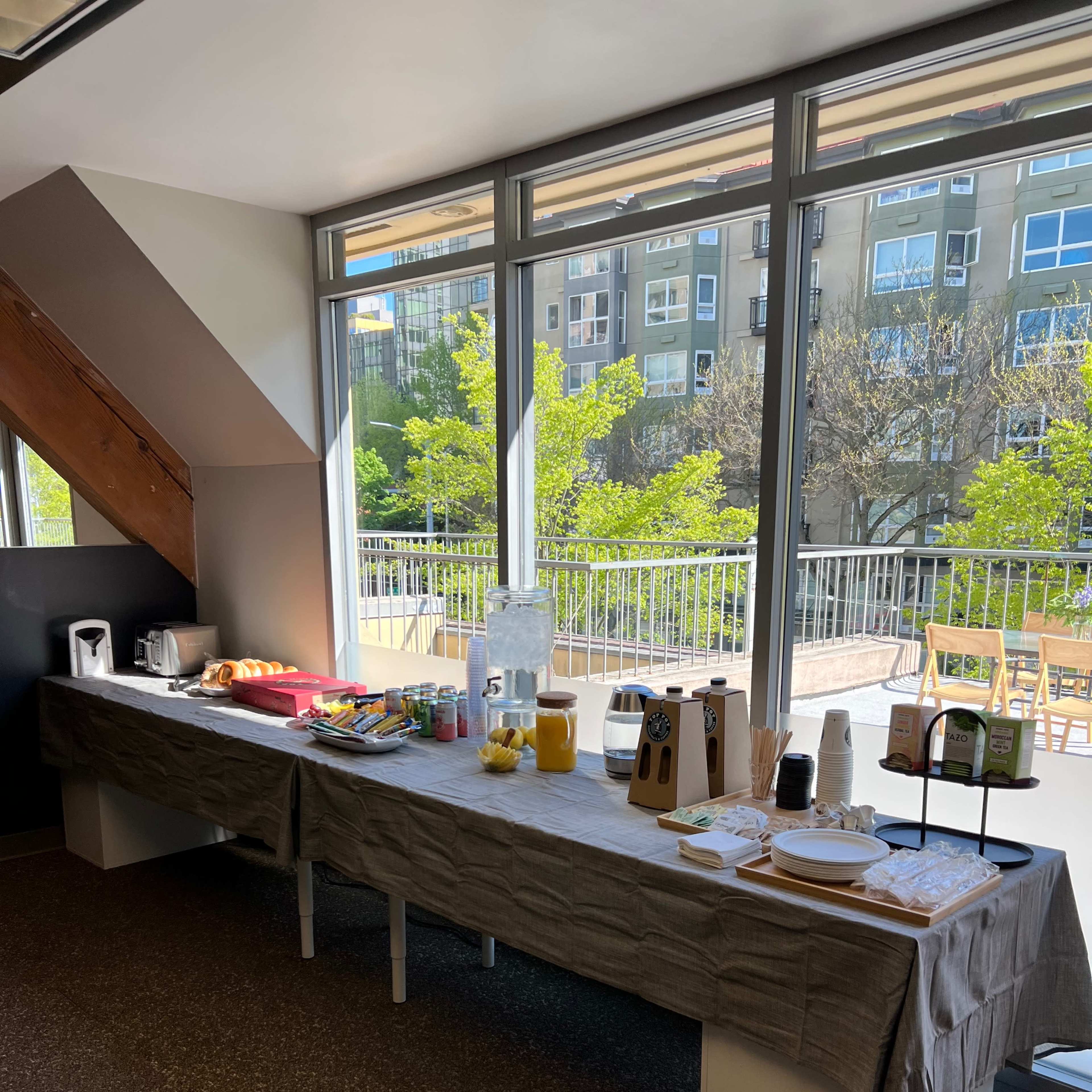 A long table is set up with food and drink items near large windows displaying greenery outside.