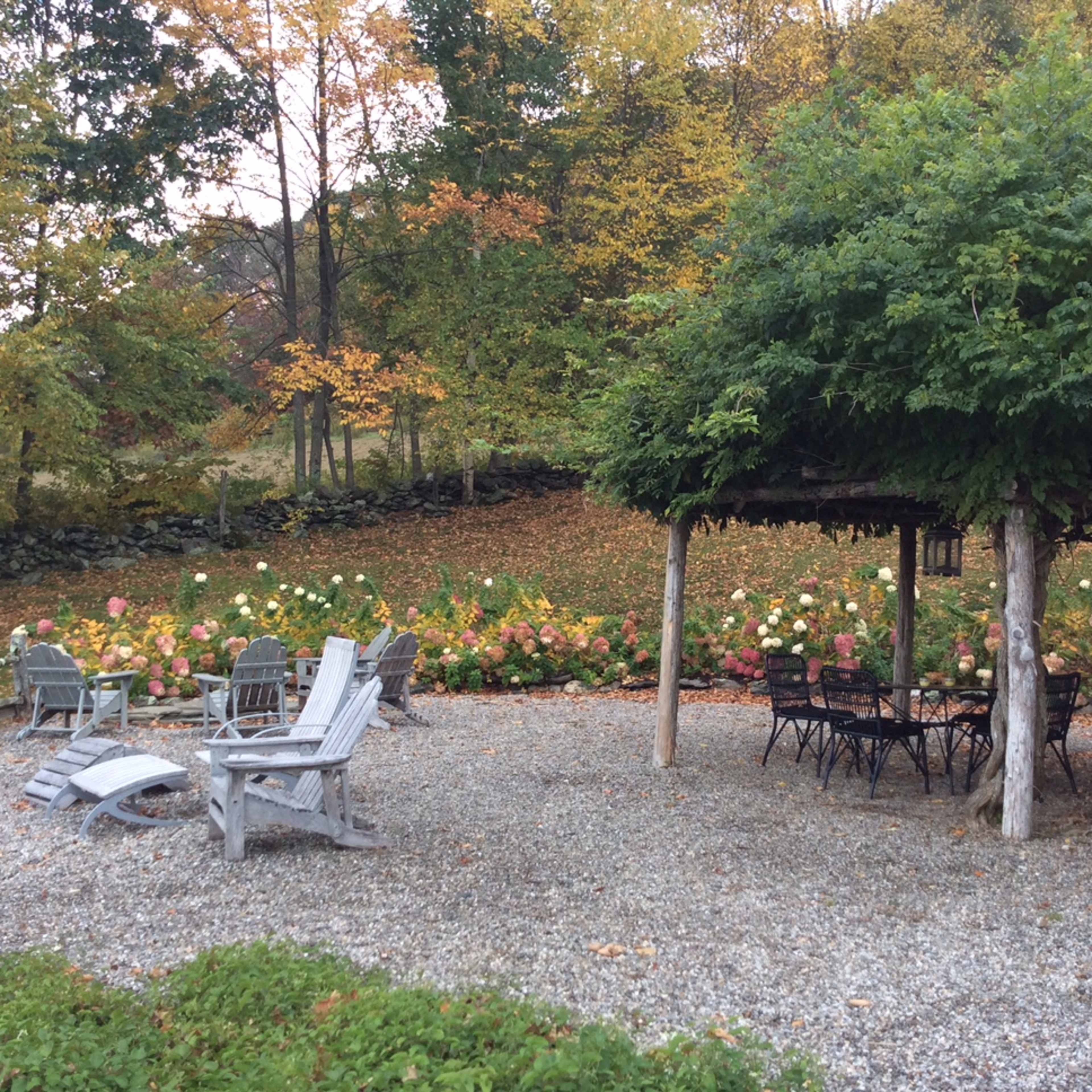 A gravel patio area with wooden chairs and a table is surrounded by blooming flowers and trees in autumn.