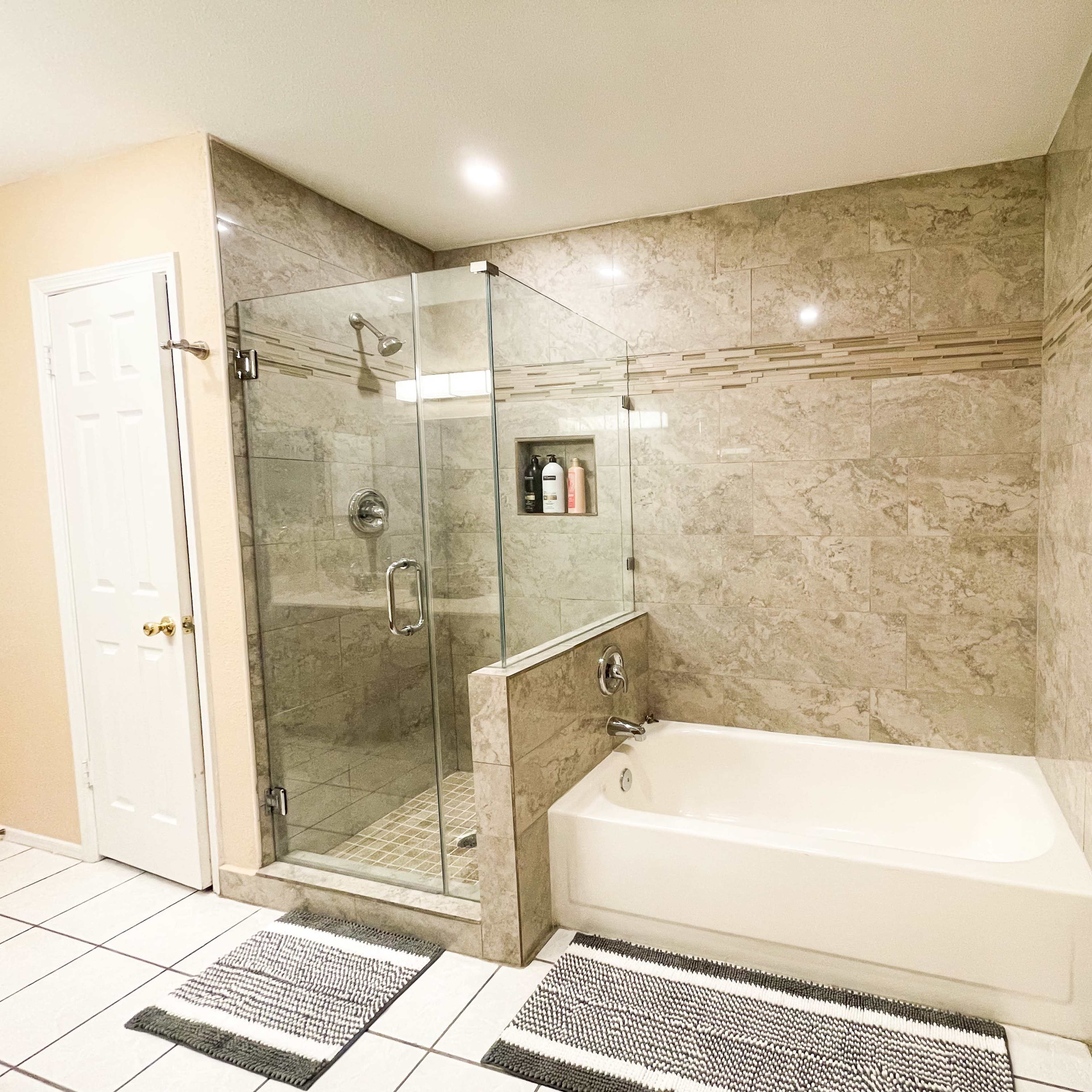 A modern bathroom featuring a glass shower enclosure, a white bathtub, and tile floors with two matching rugs.