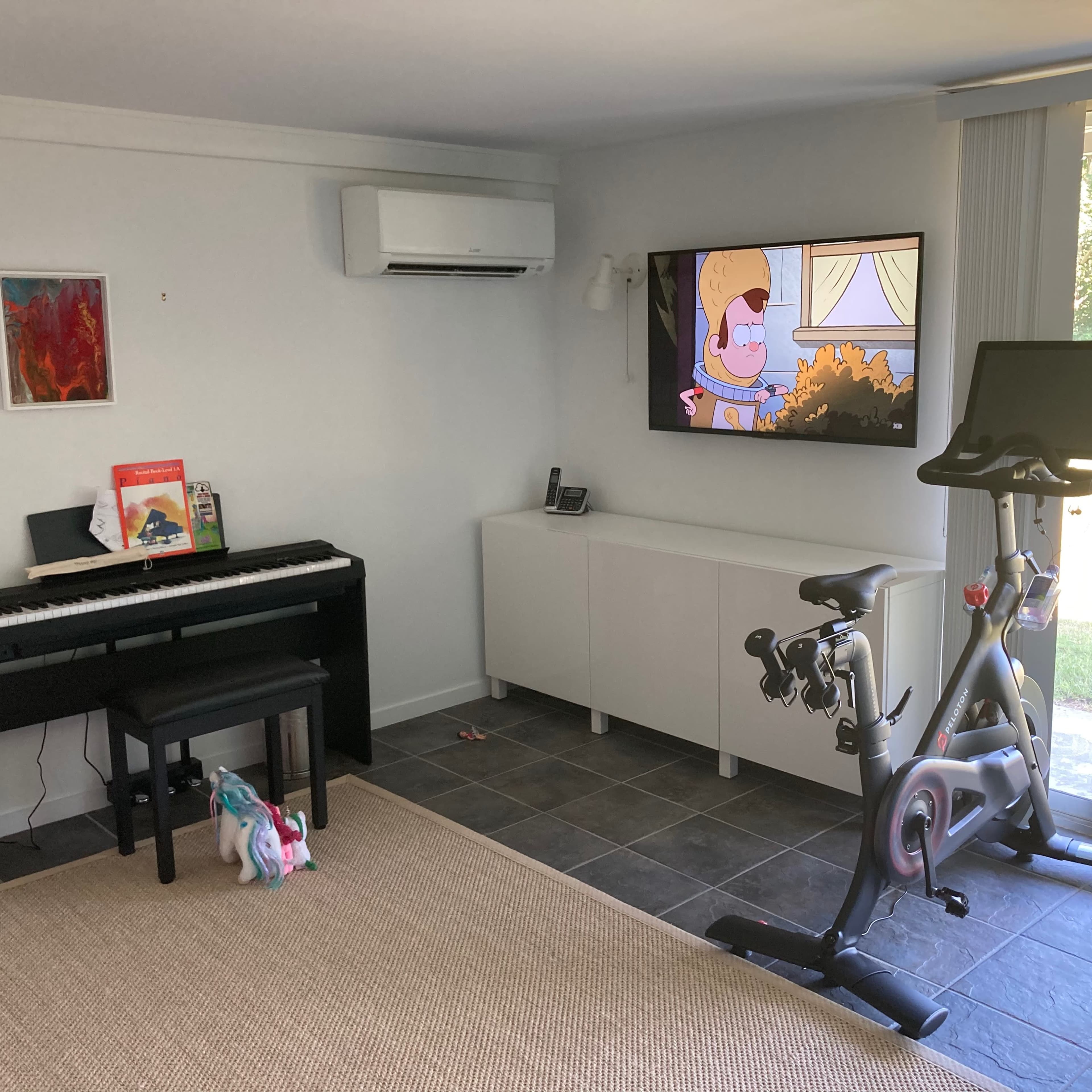 The room features a piano, a stationary bike, and a television displaying an animated show, with large windows offering a view of the outdoors.