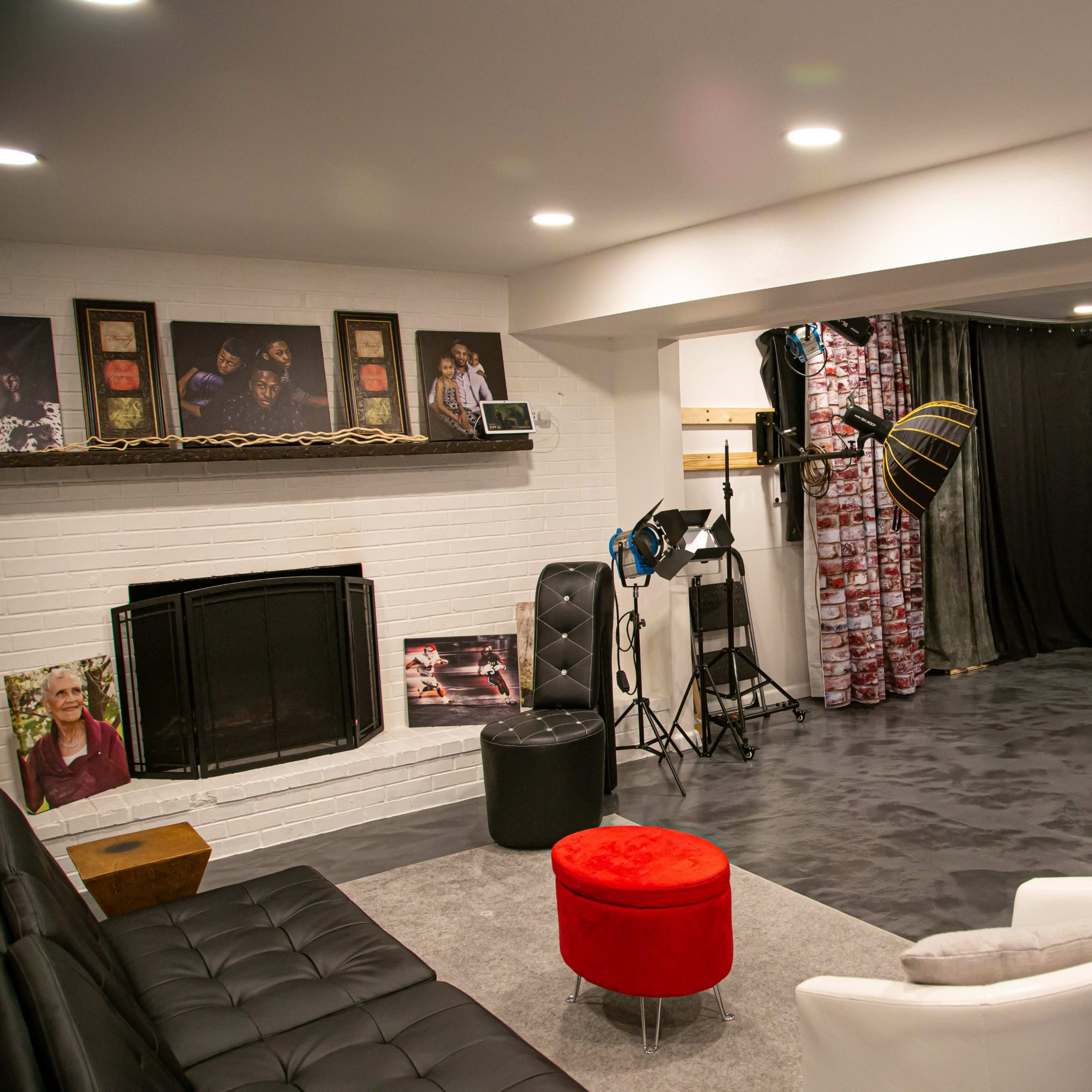 A studio space with a black backdrop, lighting equipment, a fireplace, and a seating area featuring a black couch and a red ottoman.