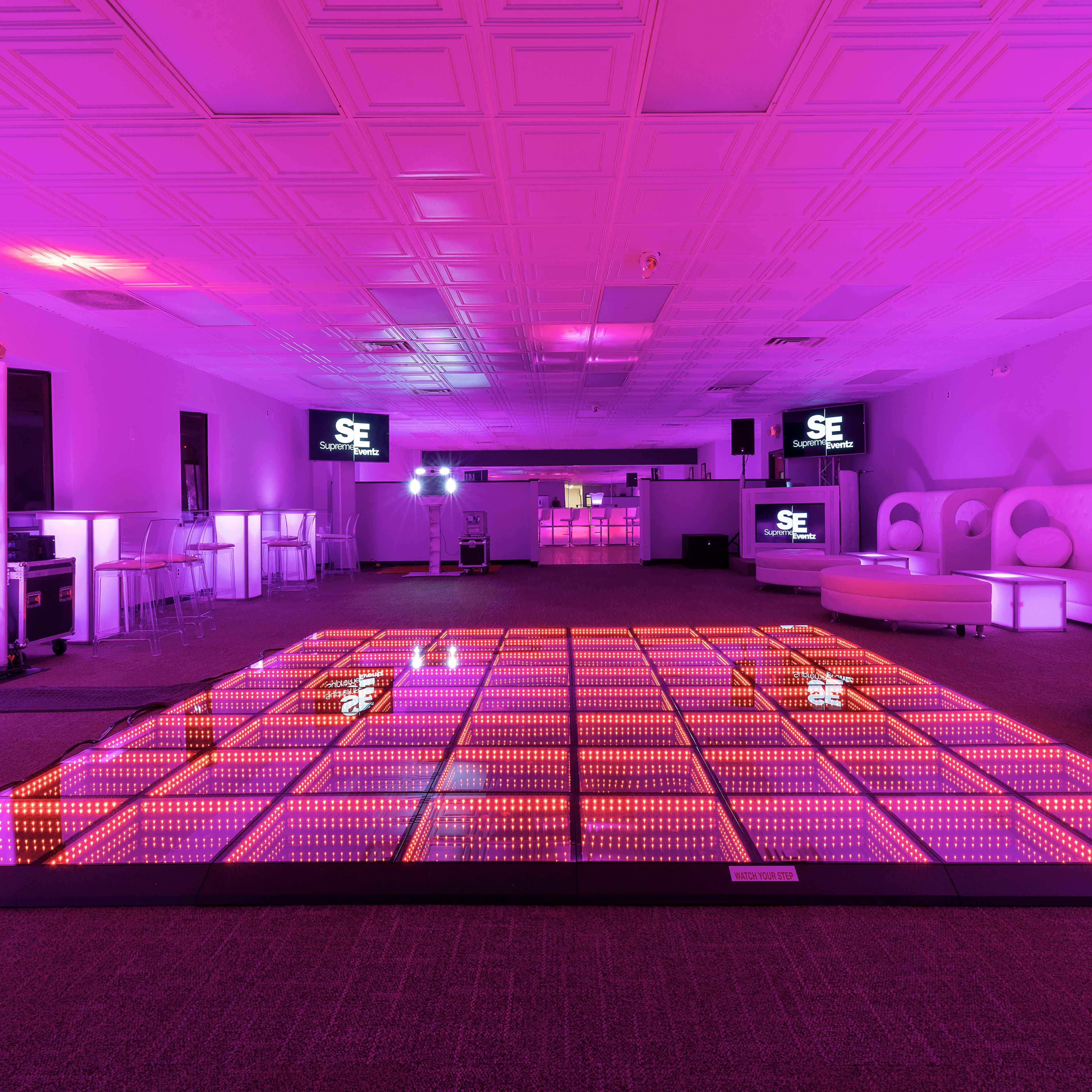 A dance floor illuminated with colorful LED lights in a spacious event venue featuring seating areas and screens.