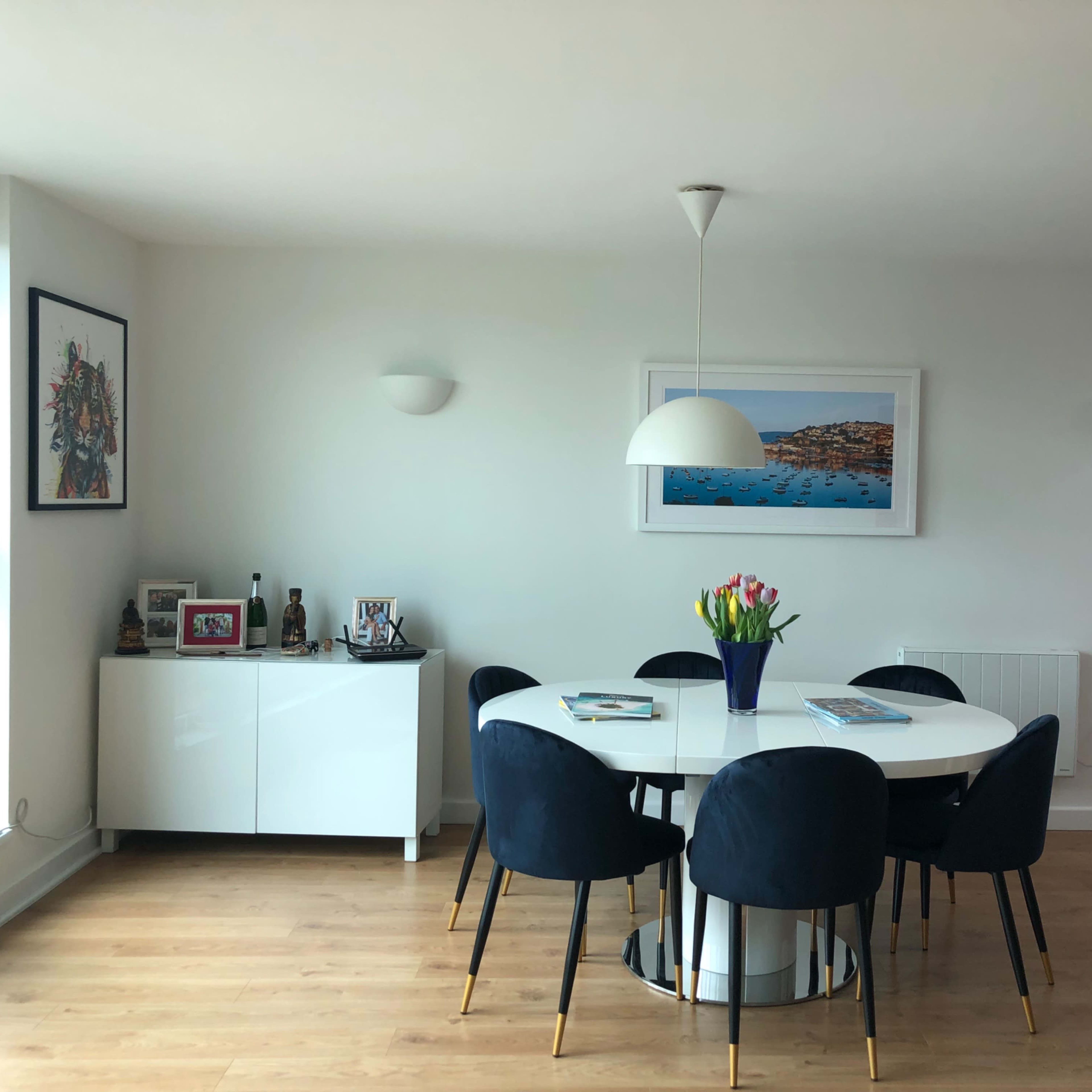 A dining area features a round white table surrounded by six black chairs, with a vase of colorful tulips and framed artwork on the walls.