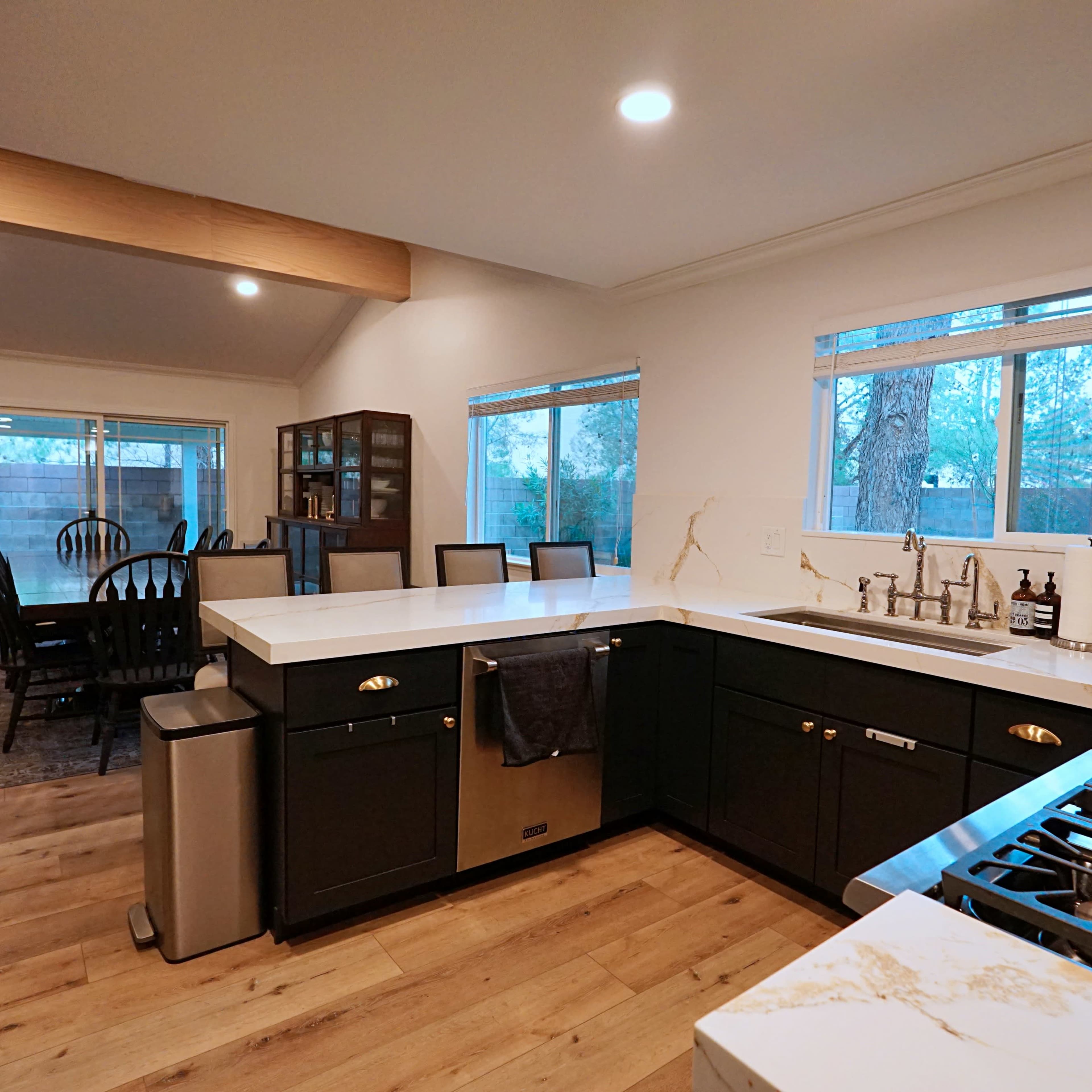 A modern kitchen features dark cabinets, a white countertop, and a stove near a large window overlooking an outdoor space.