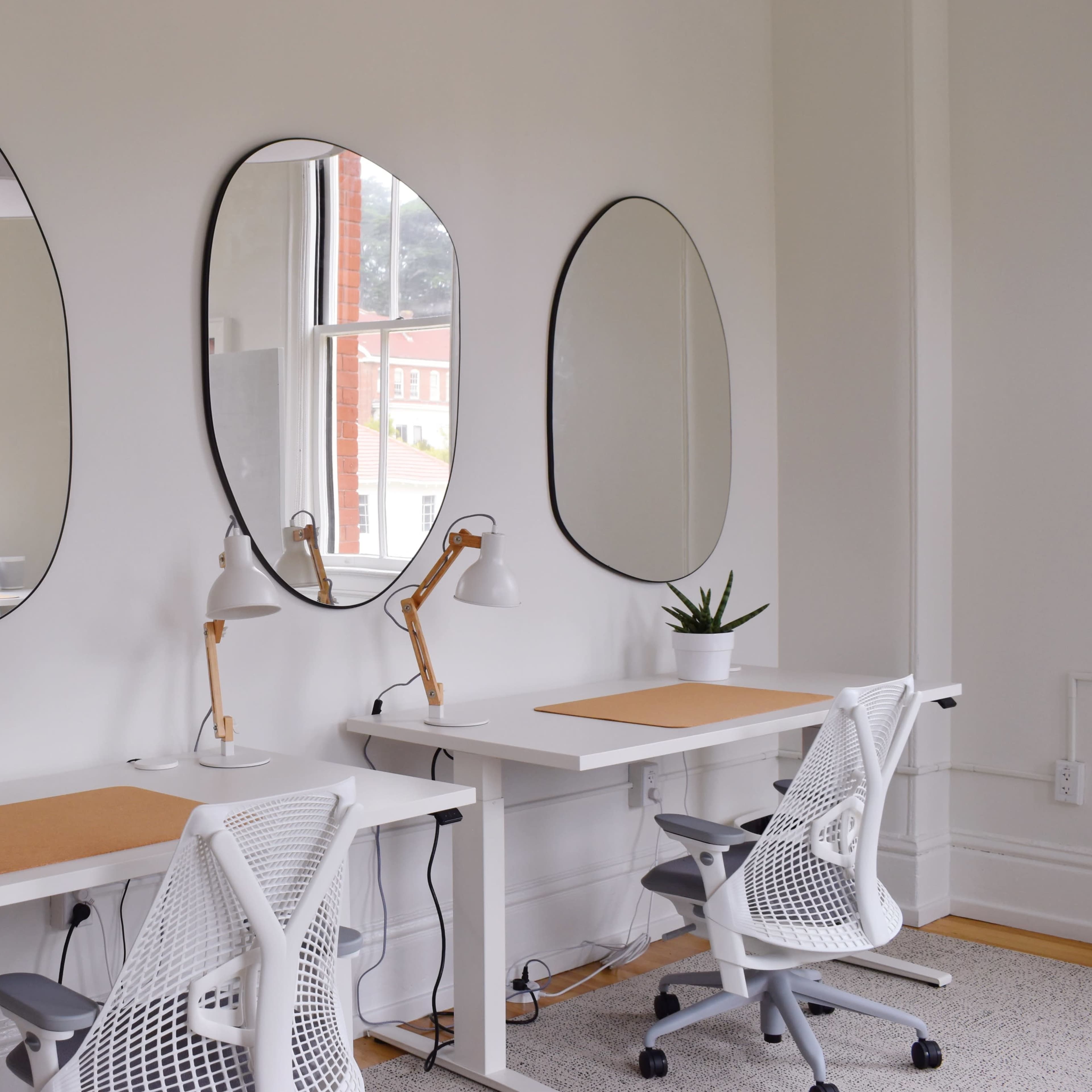 A bright workspace featuring two white desks with ergonomic chairs, three oval mirrors on the wall, and potted plants, illuminated by natural light from a window.
