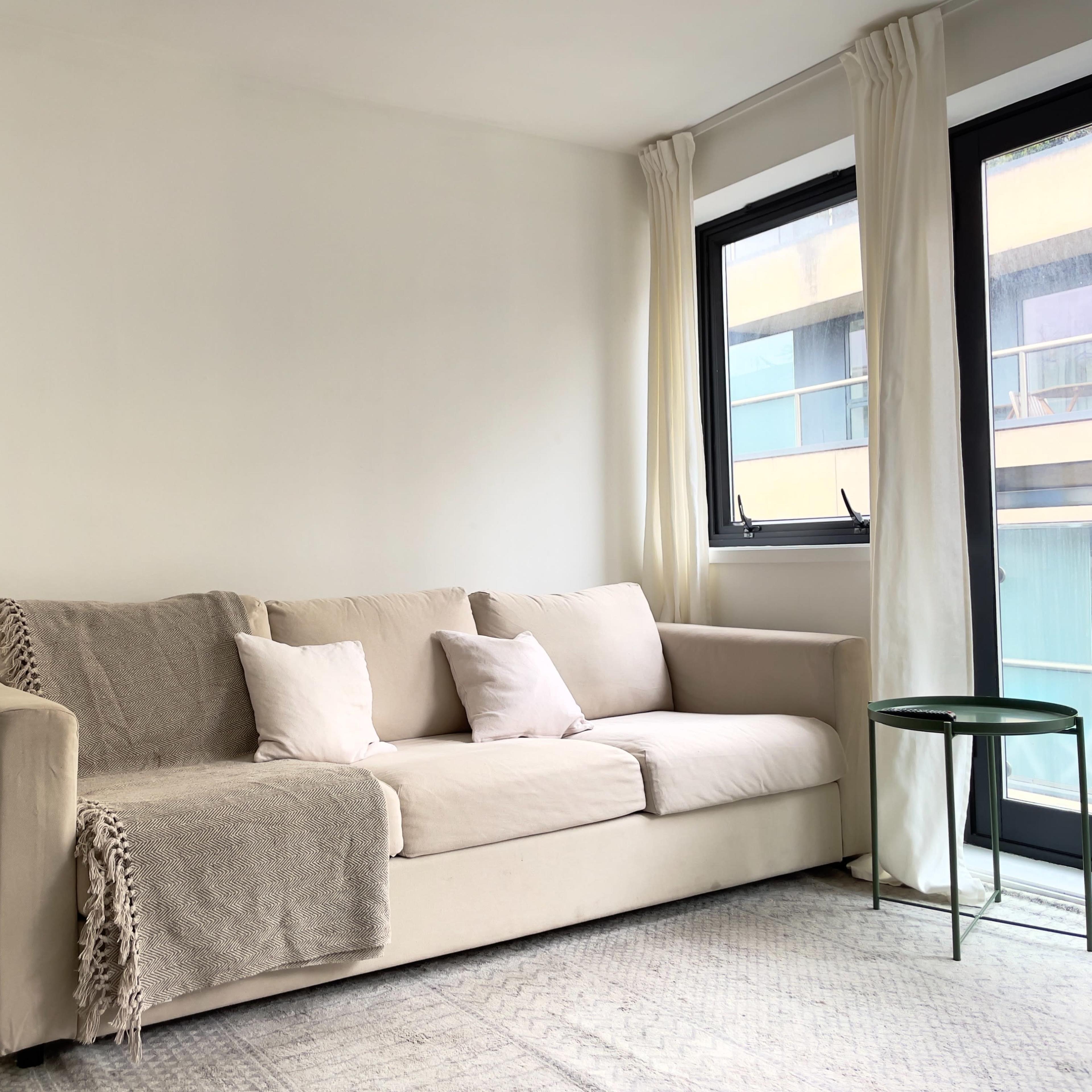 A light-colored sofa with two pillows sits against a wall next to a floor lamp and a small side table, with large windows providing natural light.