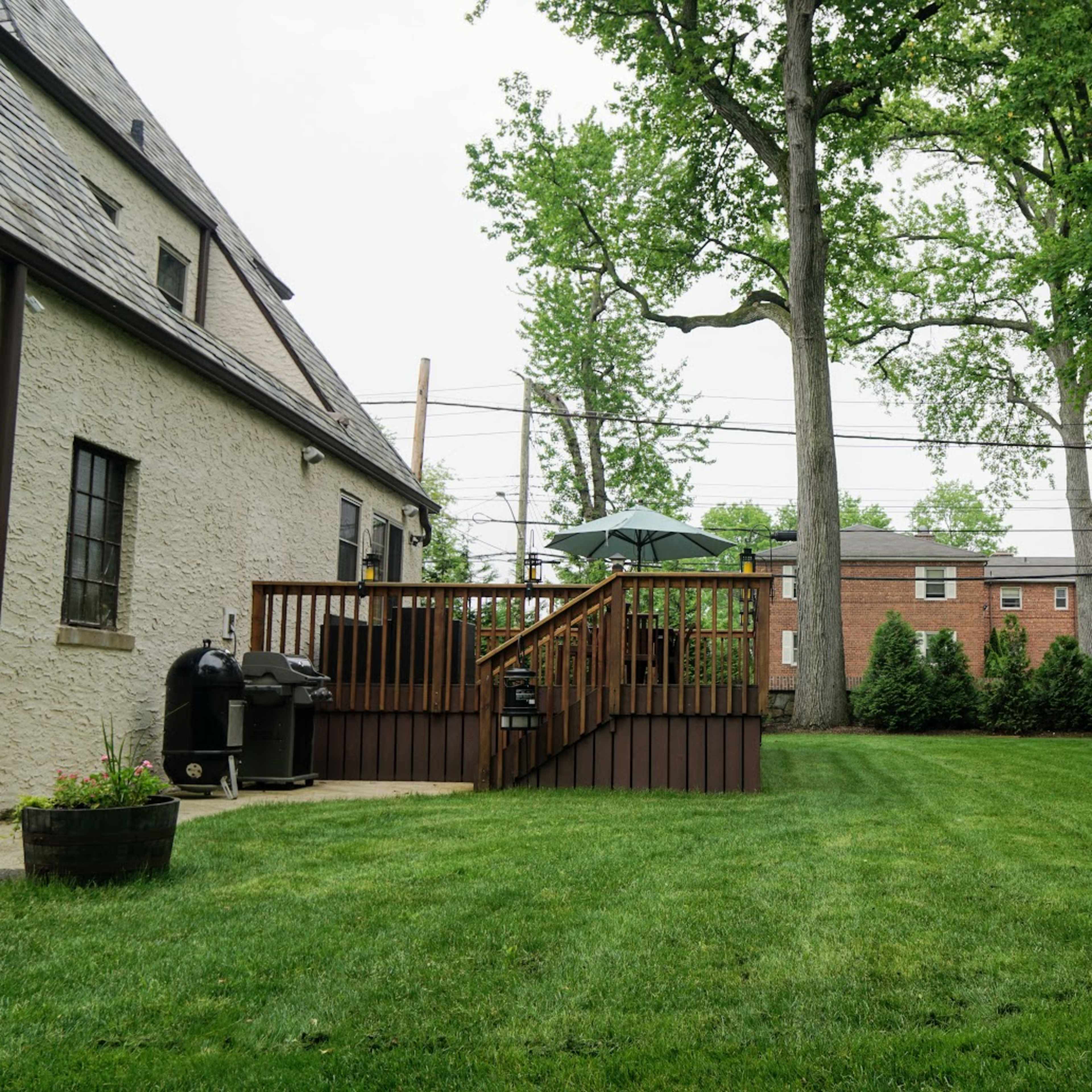 A well-maintained backyard with a wooden deck, a grill, and manicured grass, surrounded by trees and neighboring buildings.