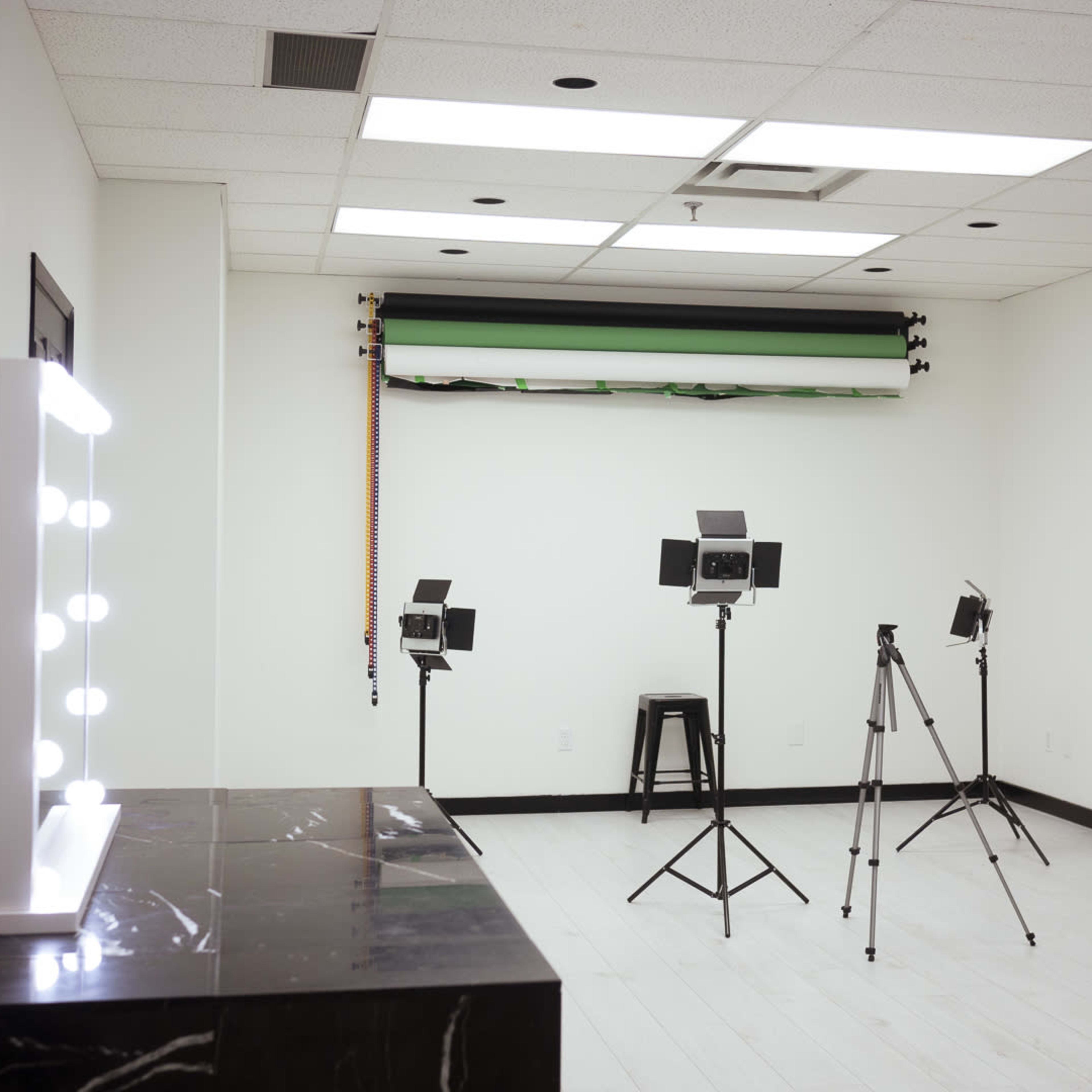 A spacious, brightly lit studio with three tripods holding lights and a vanity mirror on a dark surface, along with a backdrop system against a white wall.