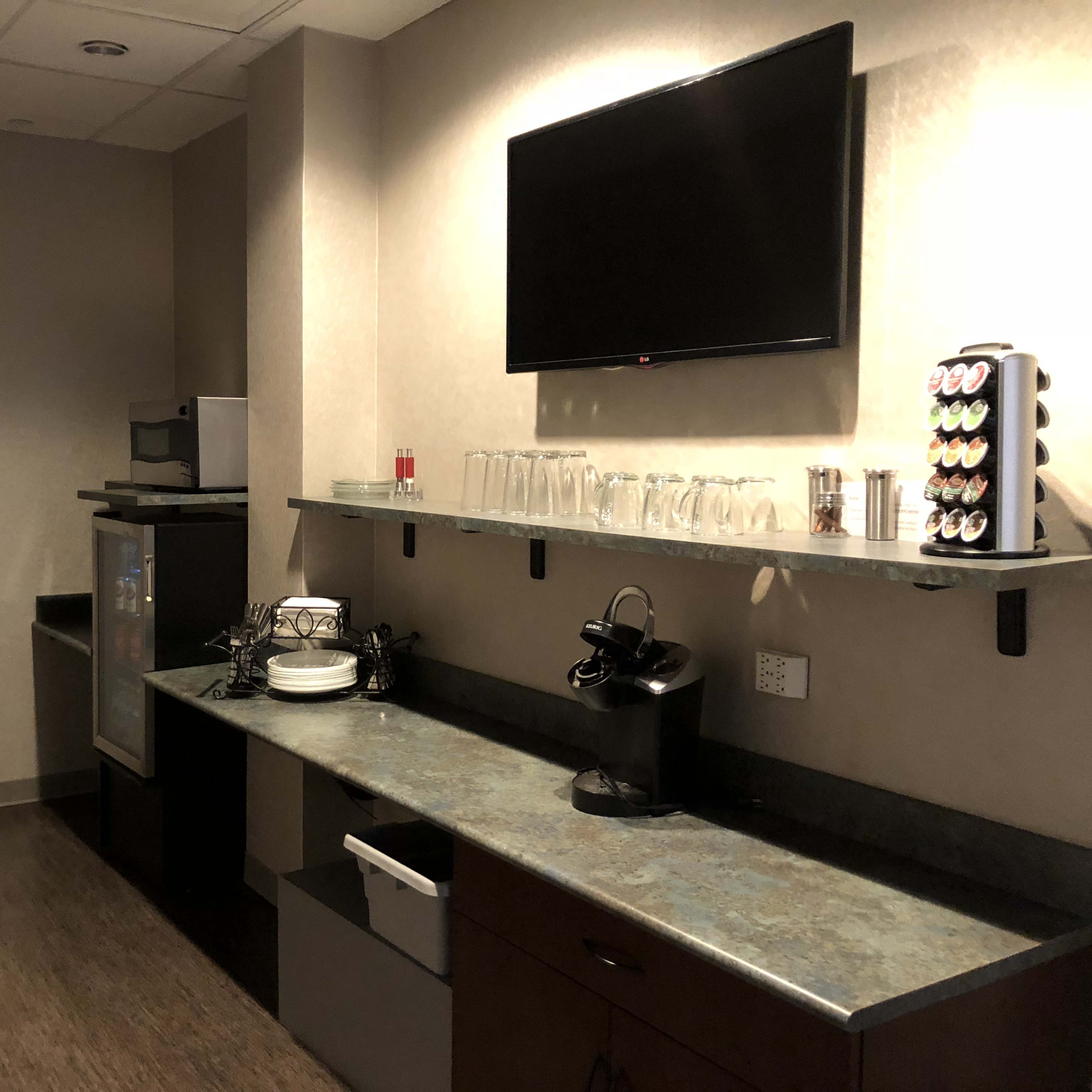 A kitchenette with a countertop, a coffee machine, glasses, and a small refrigerator against a wall, complemented by a flat-screen TV mounted above.