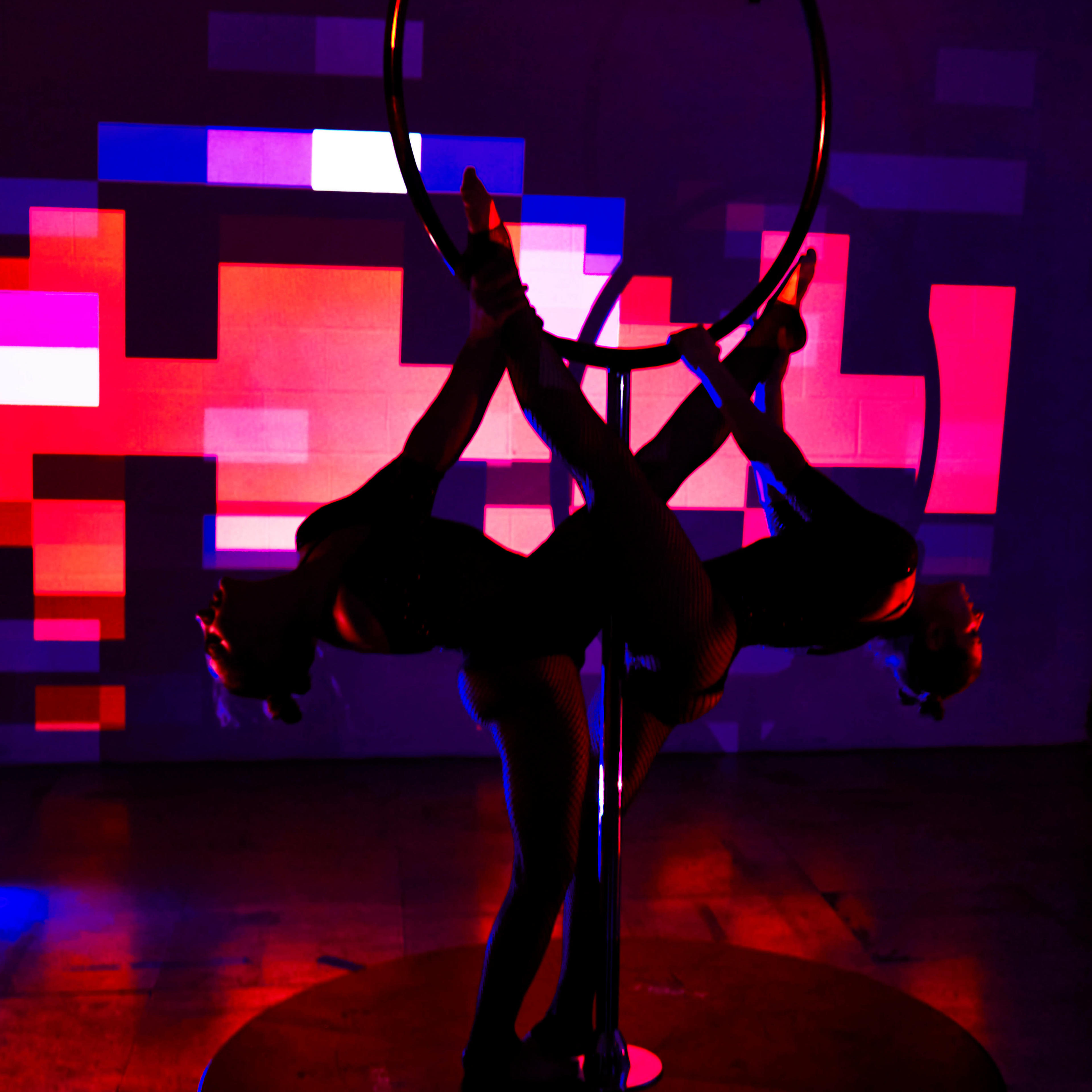Two performers in black costumes are posing on a silver aerial hoop against a backdrop of colorful, abstract lights.