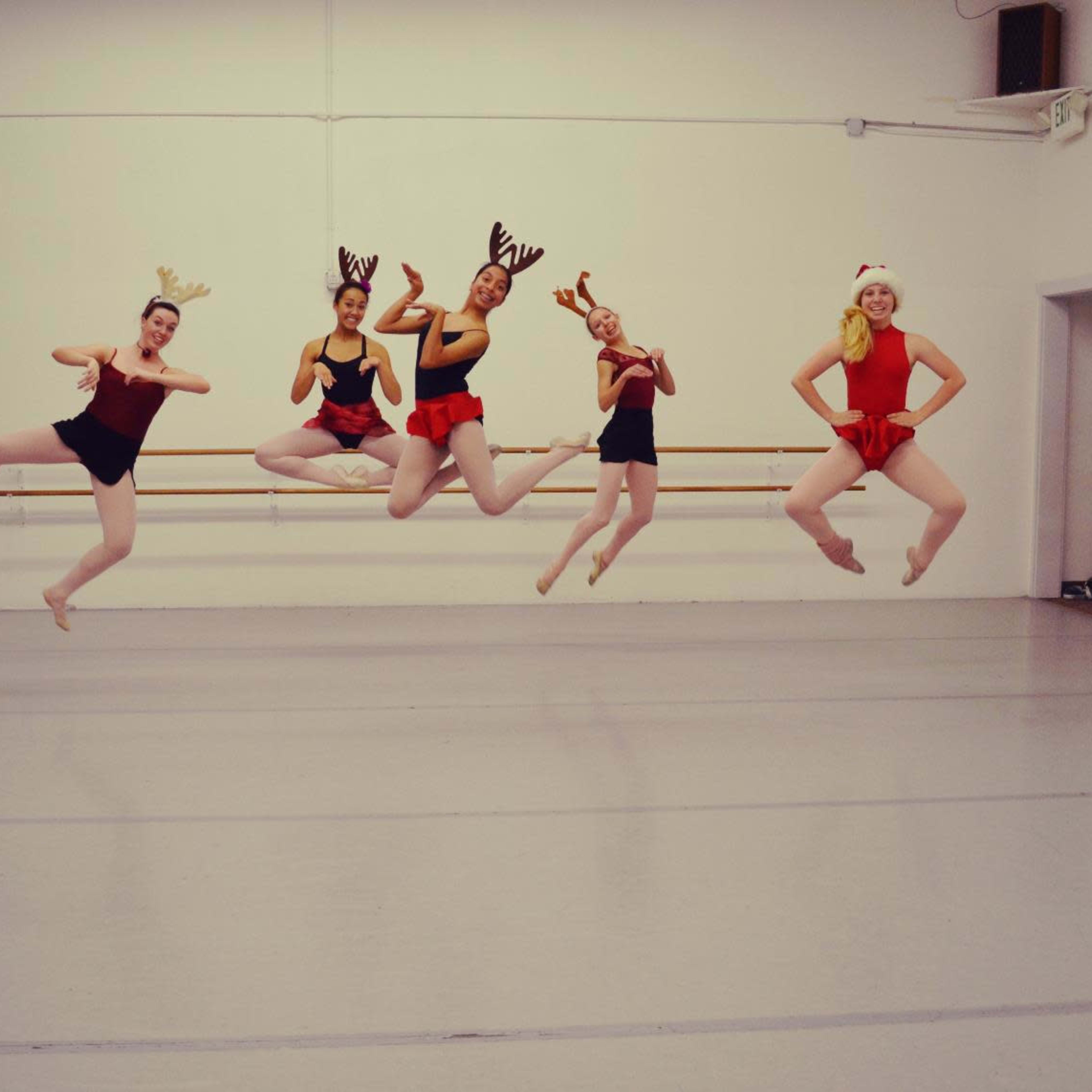 Five dancers in festive attire jump in mid-air inside a dance studio.