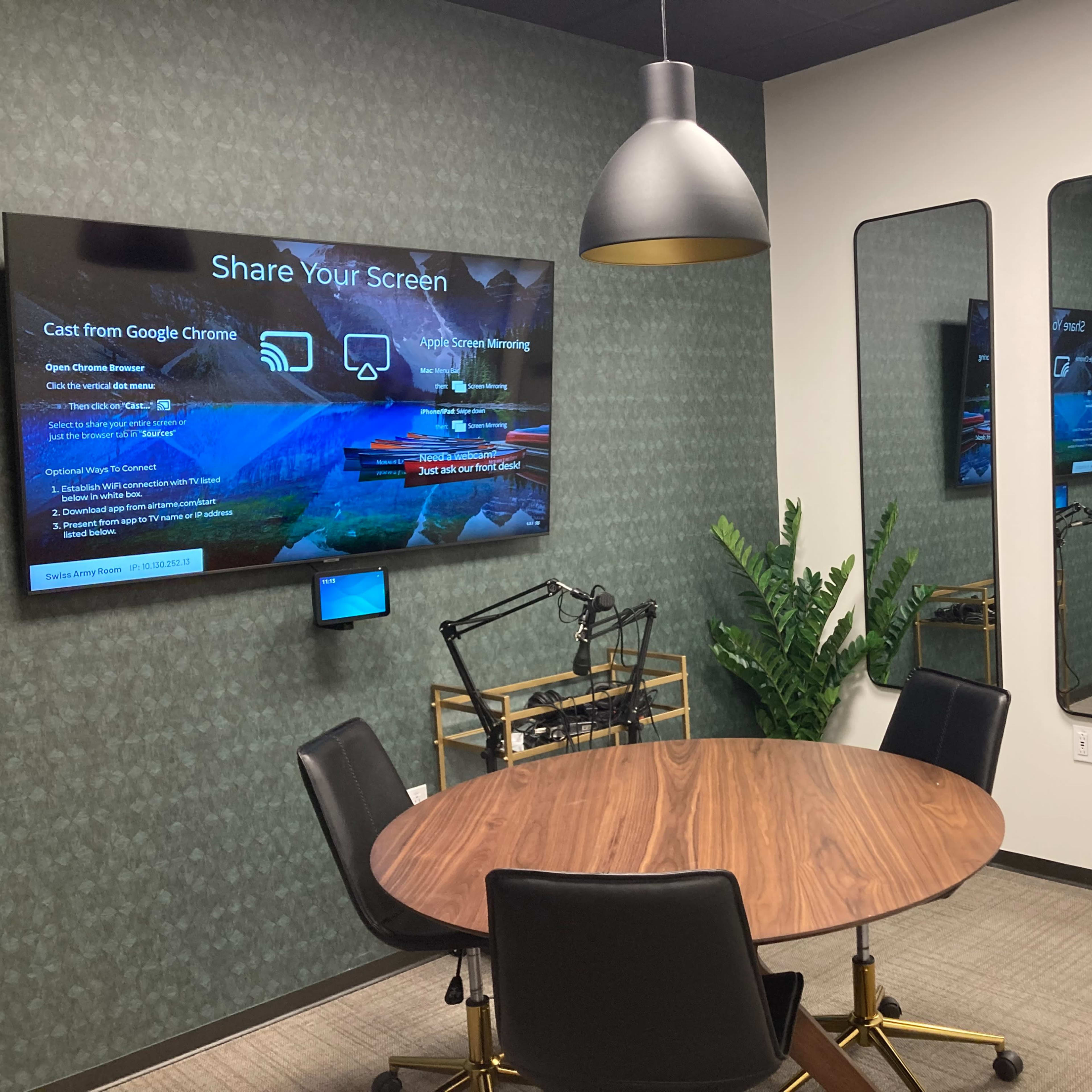 A small conference room features a round wooden table, black chairs, and two displays on the walls, one of which provides instructions for screen sharing.