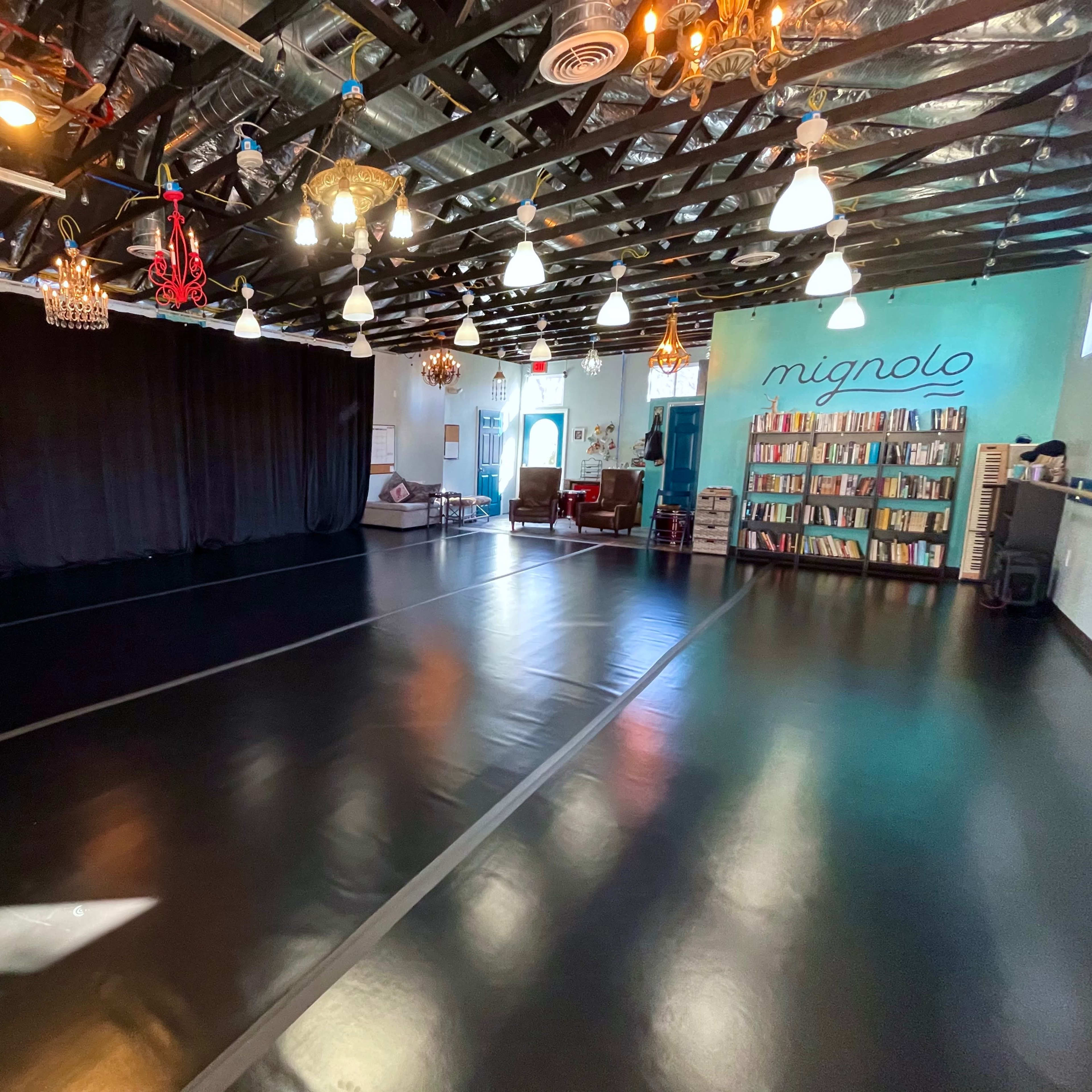 A spacious studio with black flooring, a large black curtain on one side, and a bookshelf filled with books against a wall on the other side, illuminated by various hanging lights.