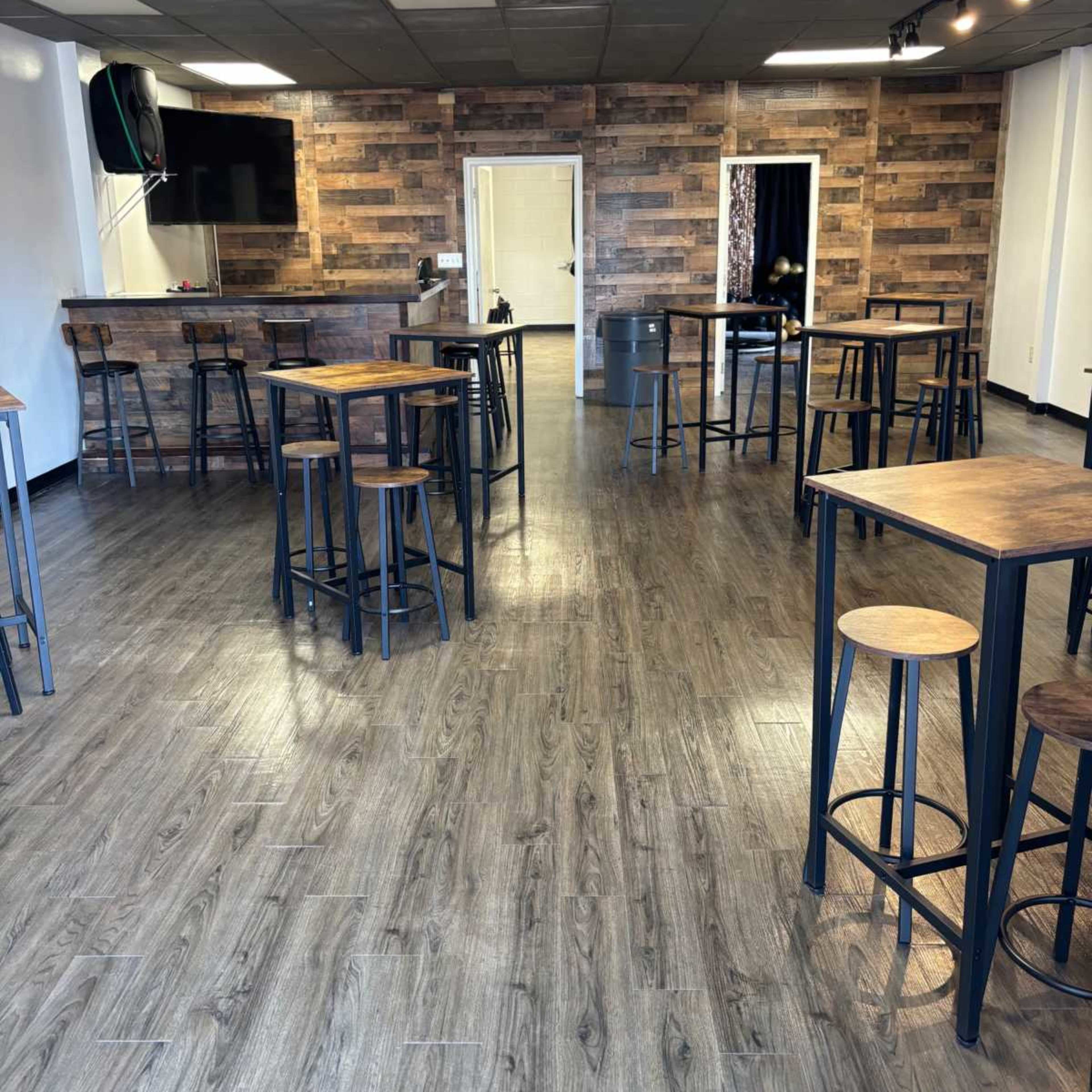 The interior of a spacious venue features several tall tables and stools arranged on a wooden floor, with a rustic wood-paneled wall in the background.