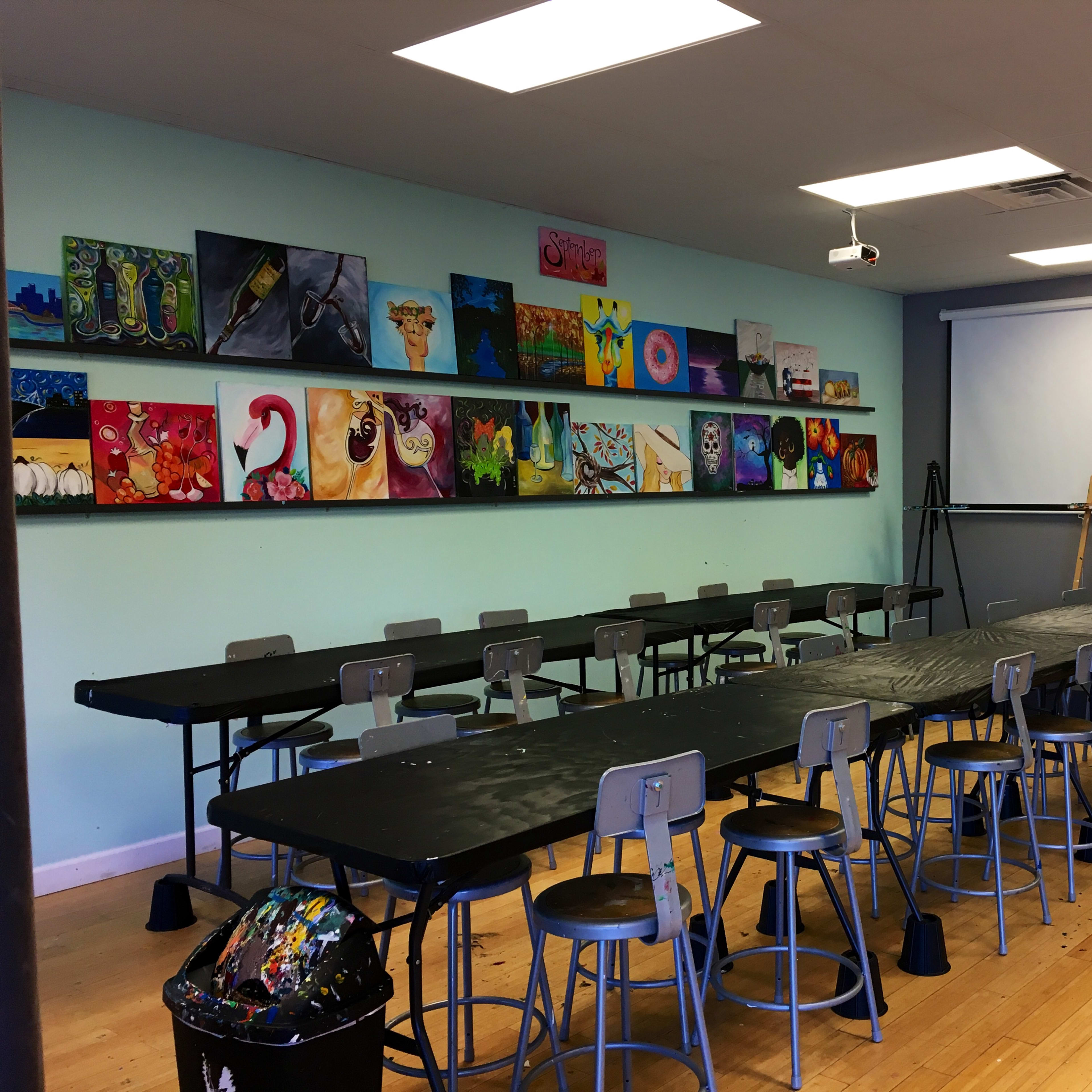 A spacious art classroom features long black tables and metal chairs, with colorful paintings displayed on the wall above and art supplies in the corner.