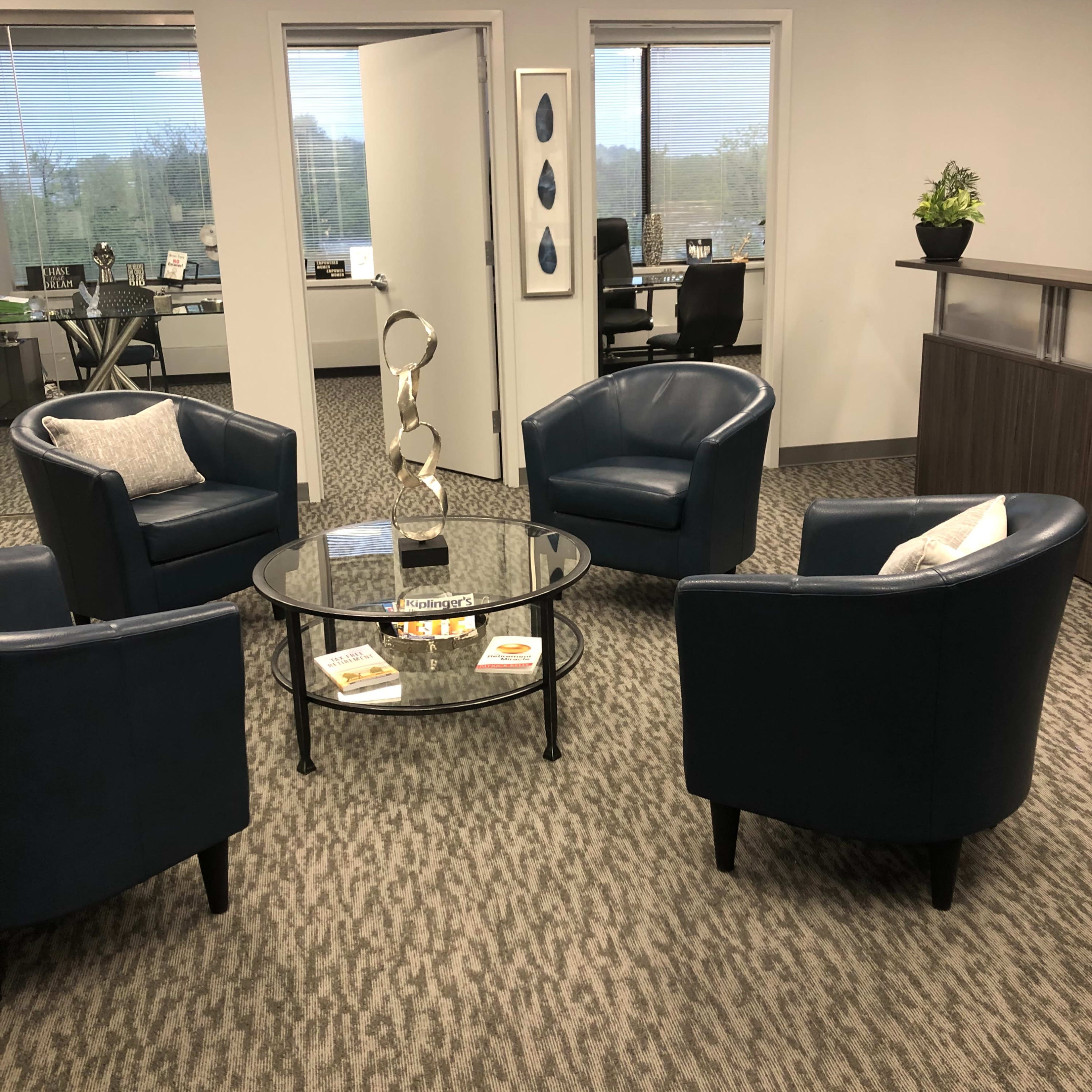 A modern office reception area with four blue armchairs arranged around a glass coffee table on a patterned carpet.