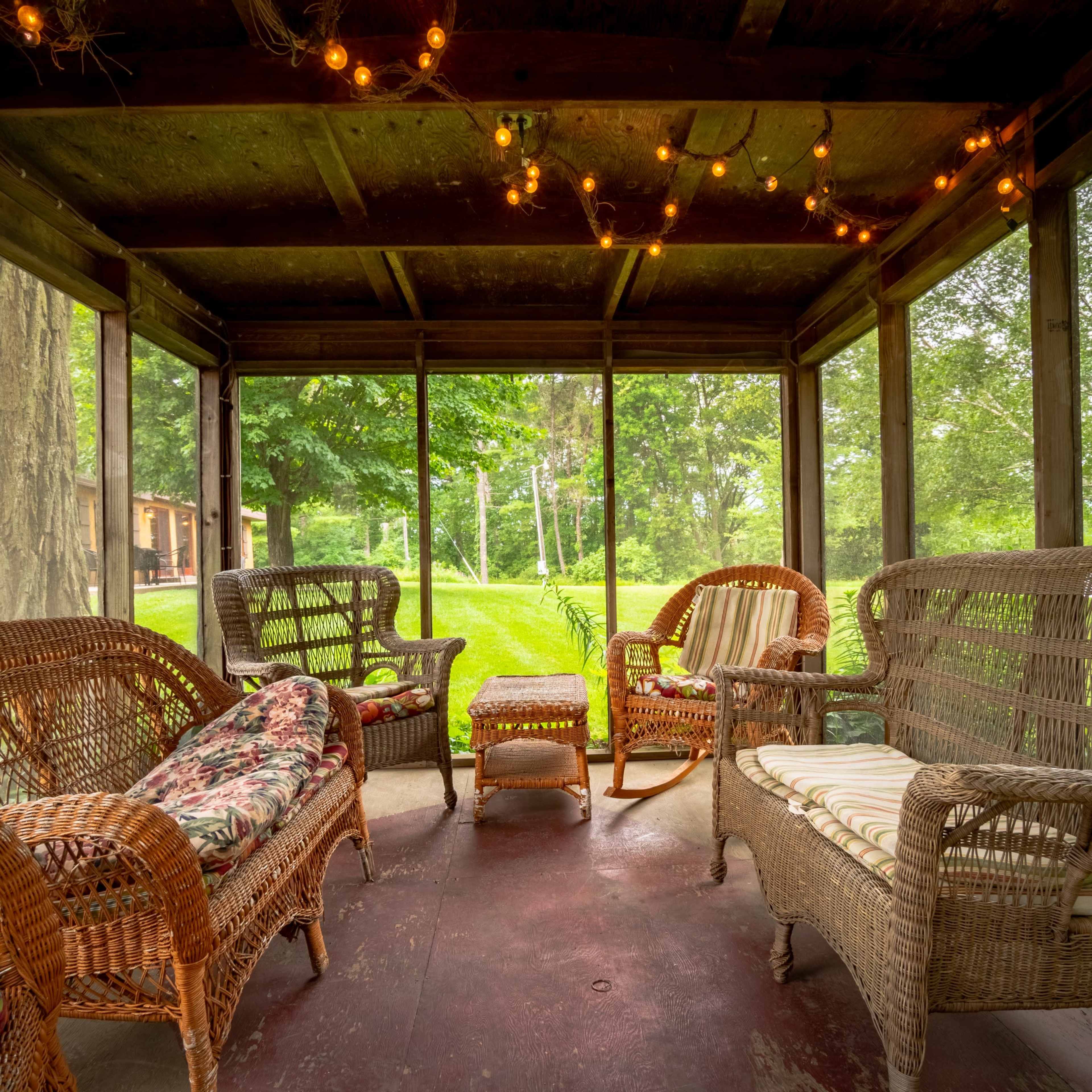 A screened porch furnished with several wicker chairs, a small table, and surrounded by greenery.