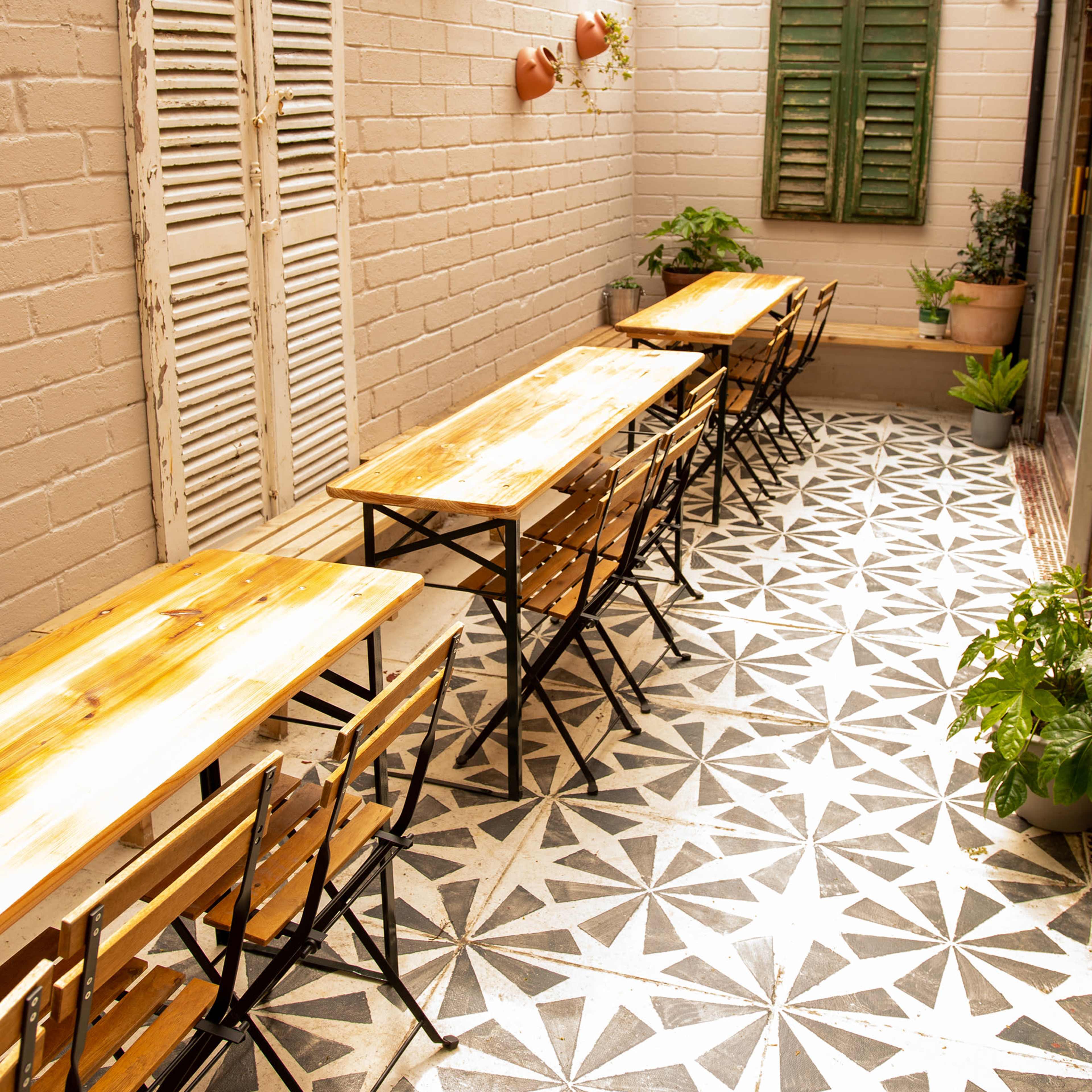 A cozy outdoor seating area features wooden tables and chairs arranged on a patterned tiled floor beneath a wooden pergola adorned with plants.