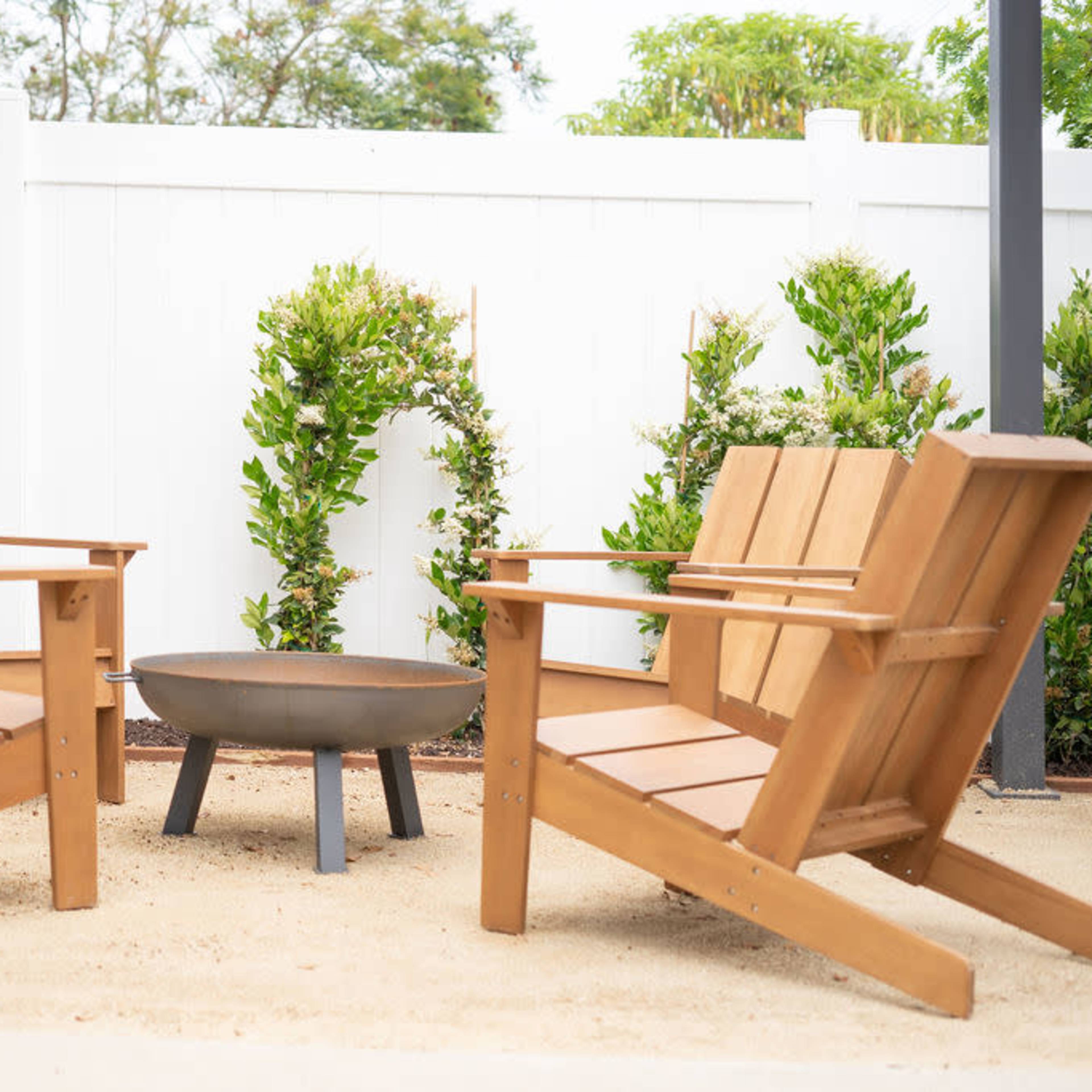 Two wooden Adirondack chairs are positioned facing a round fire pit, surrounded by a sandy area and small greenery against a white fence.
