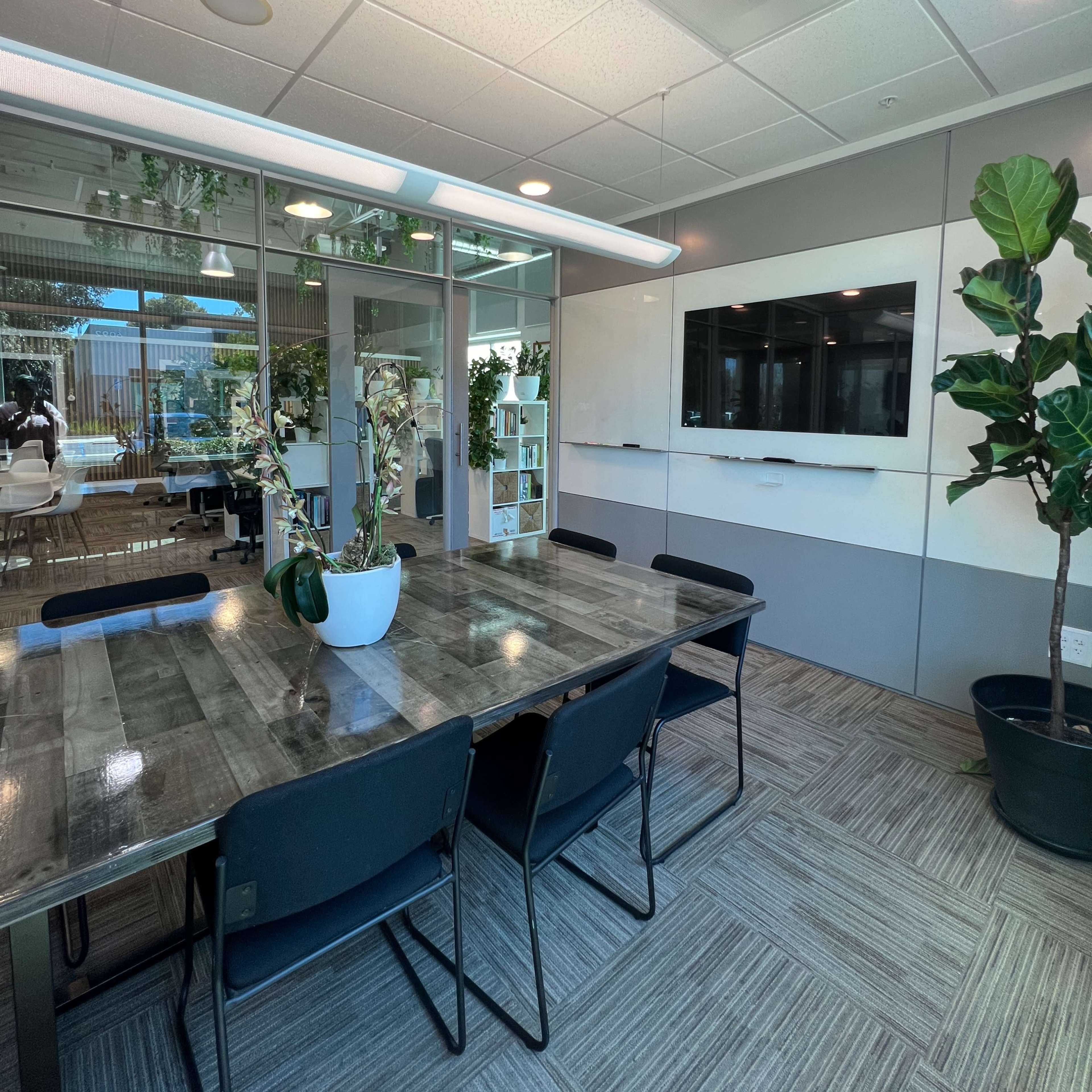 A modern conference room features a large wooden table surrounded by black chairs, with plants and a wall-mounted screen in the background.