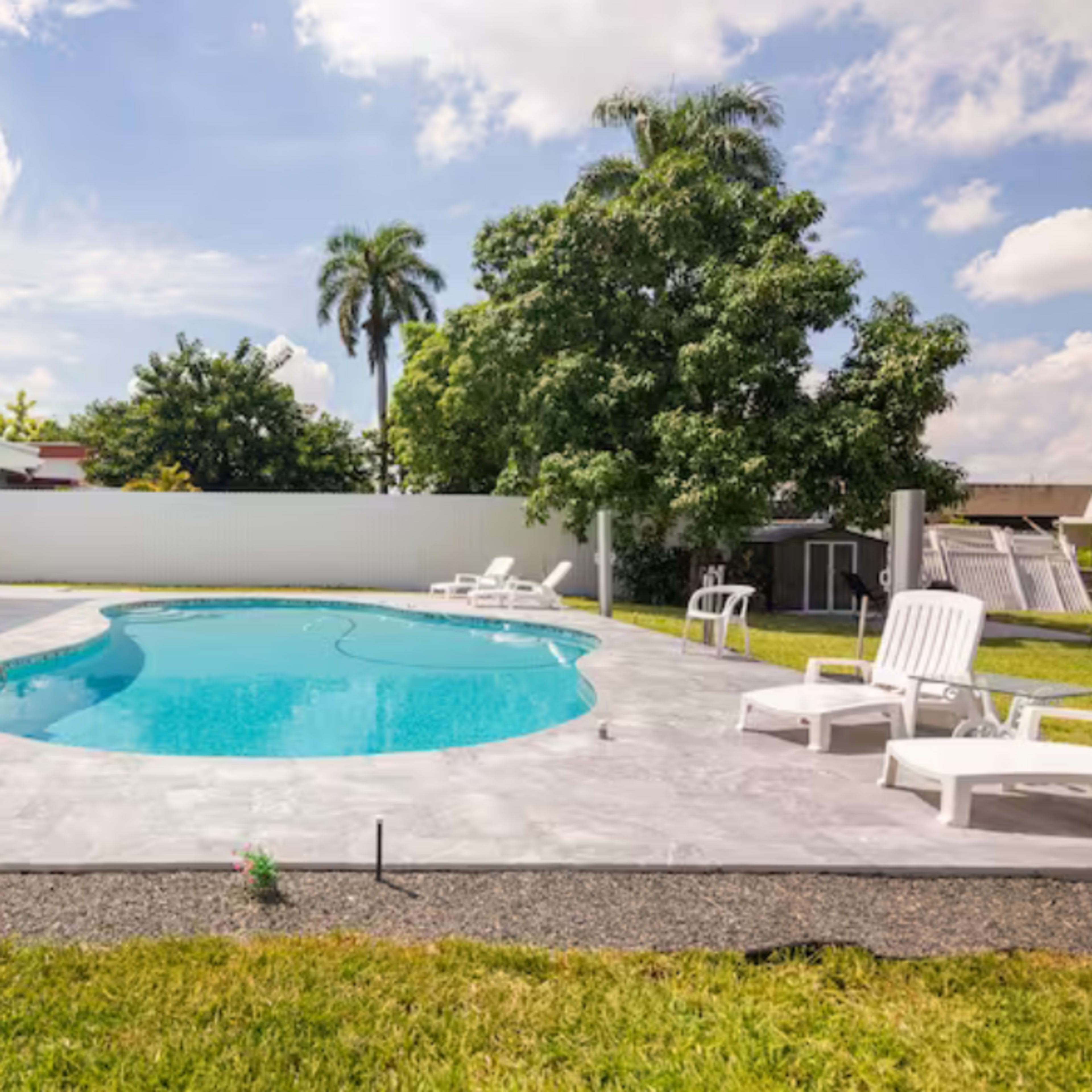 A bright outdoor space features a swimming pool surrounded by lounge chairs and green grass, with a house and palm trees in the background.