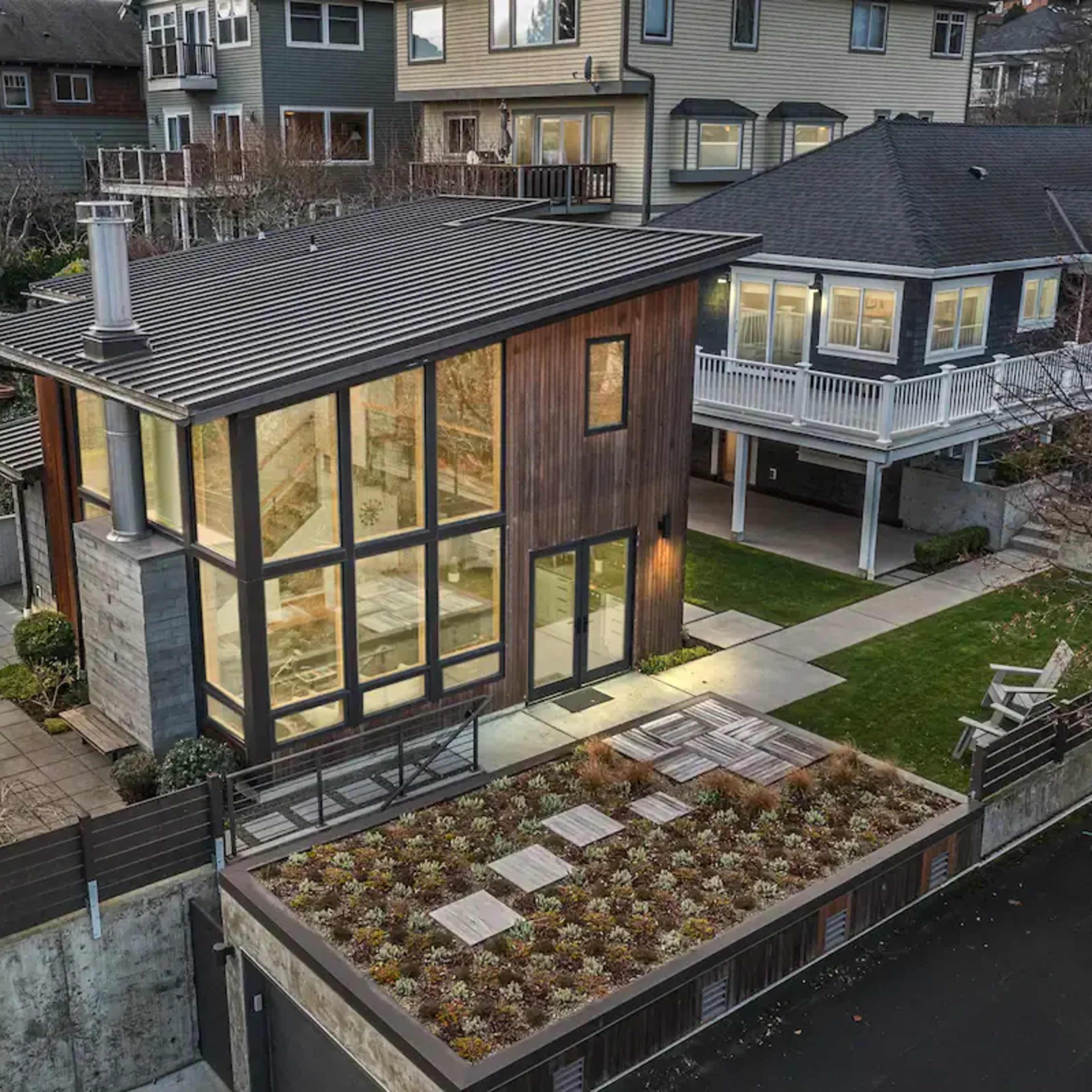A modern two-story house with large glass windows and wooden siding is situated next to traditional homes, surrounded by landscaped gardens.