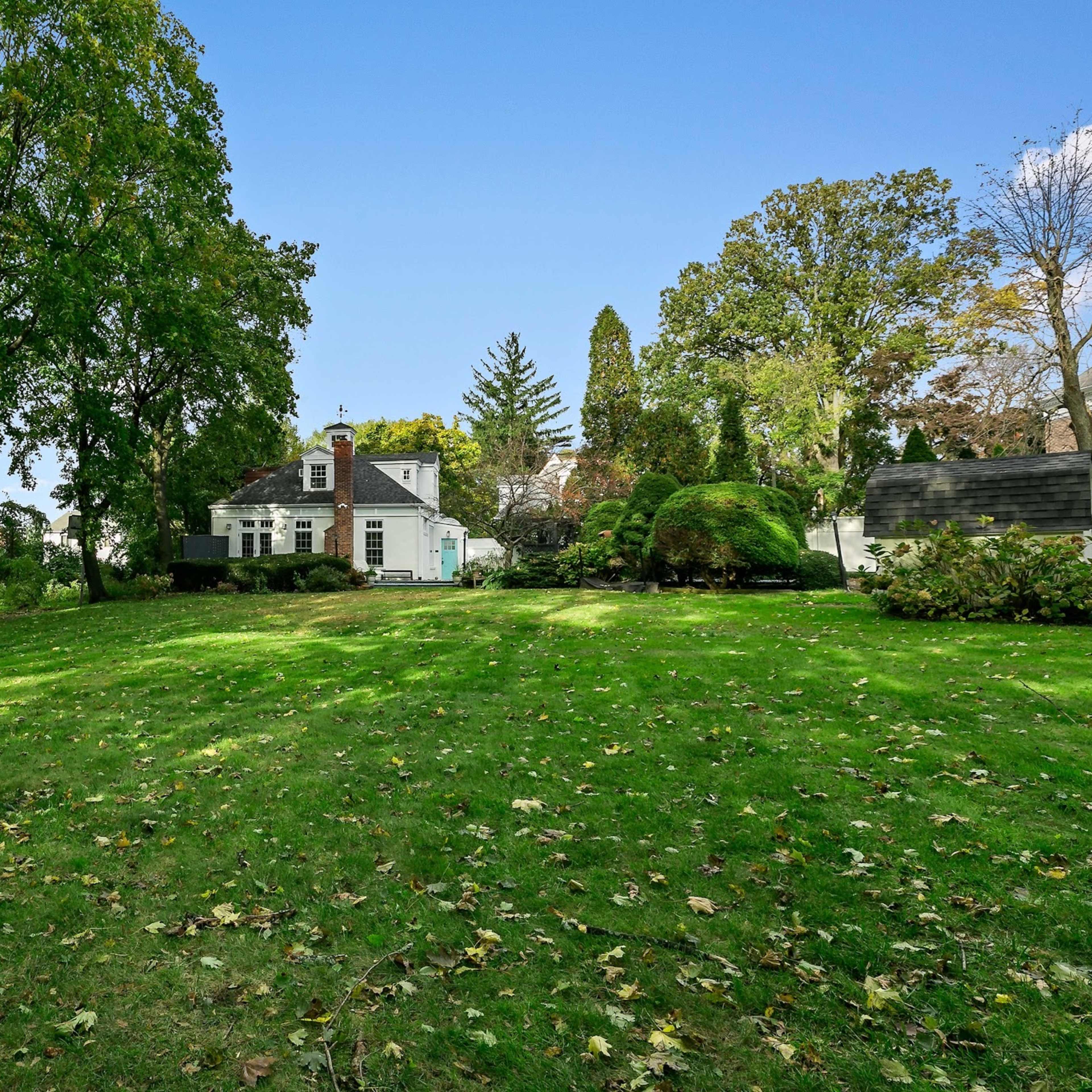 A house with a sloped lawn and several trees is situated in a residential area under a clear blue sky.