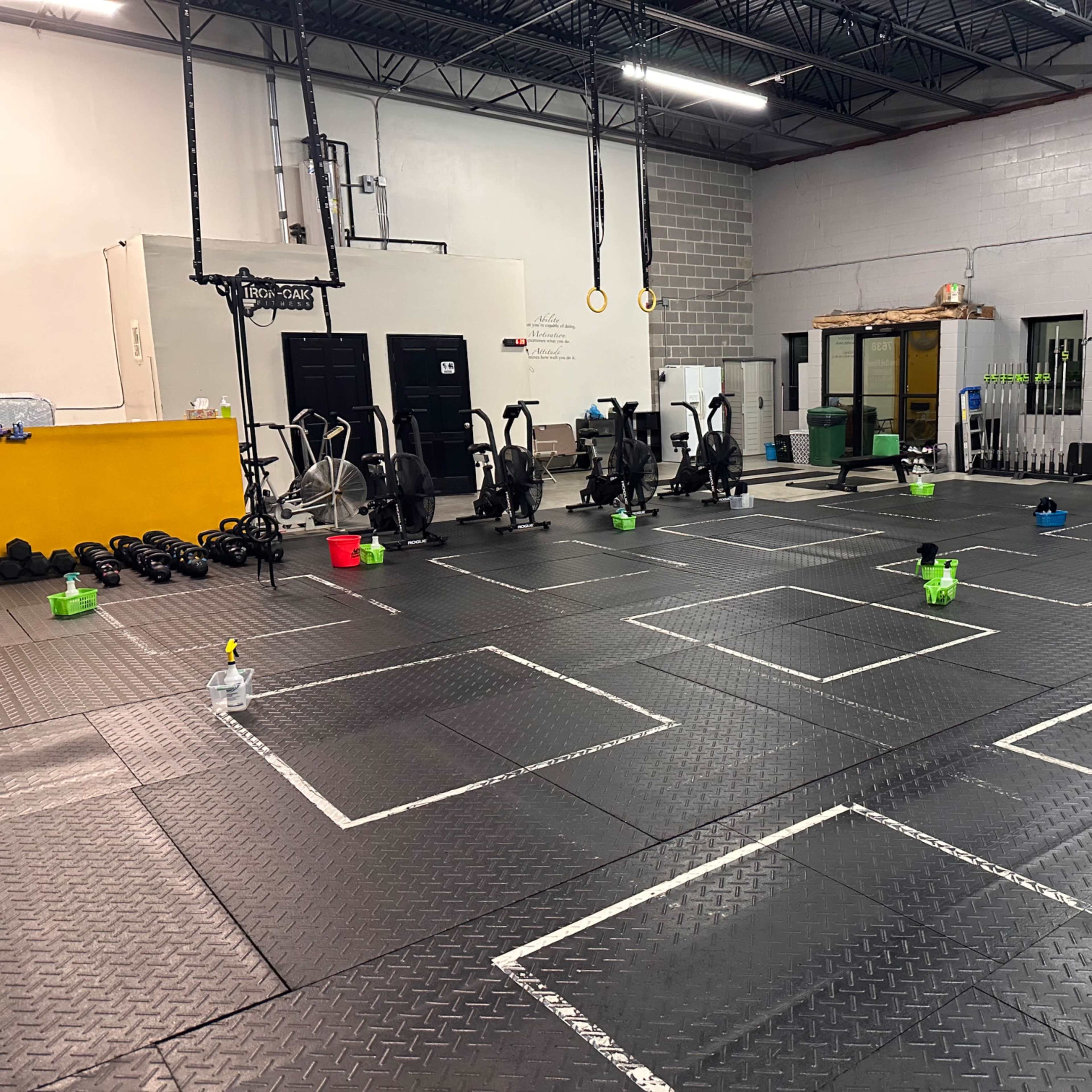 A spacious gym with exercise equipment, including stationary bikes and weights, arranged in sections defined by white tape on a black rubber floor.