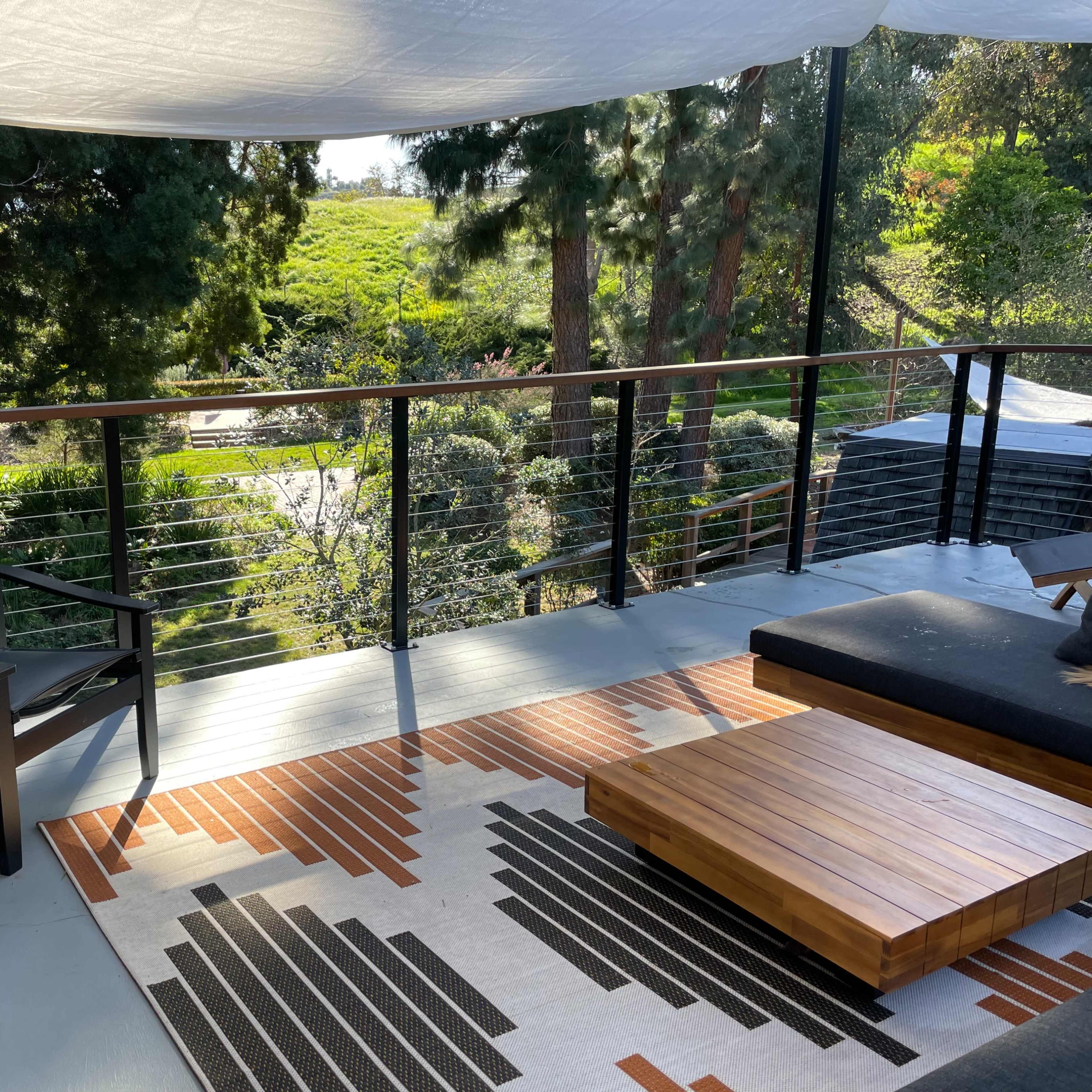 A modern outdoor deck features a sleek seating area with a black chair, a wooden coffee table, and a patterned rug, overlooking a lush green landscape.