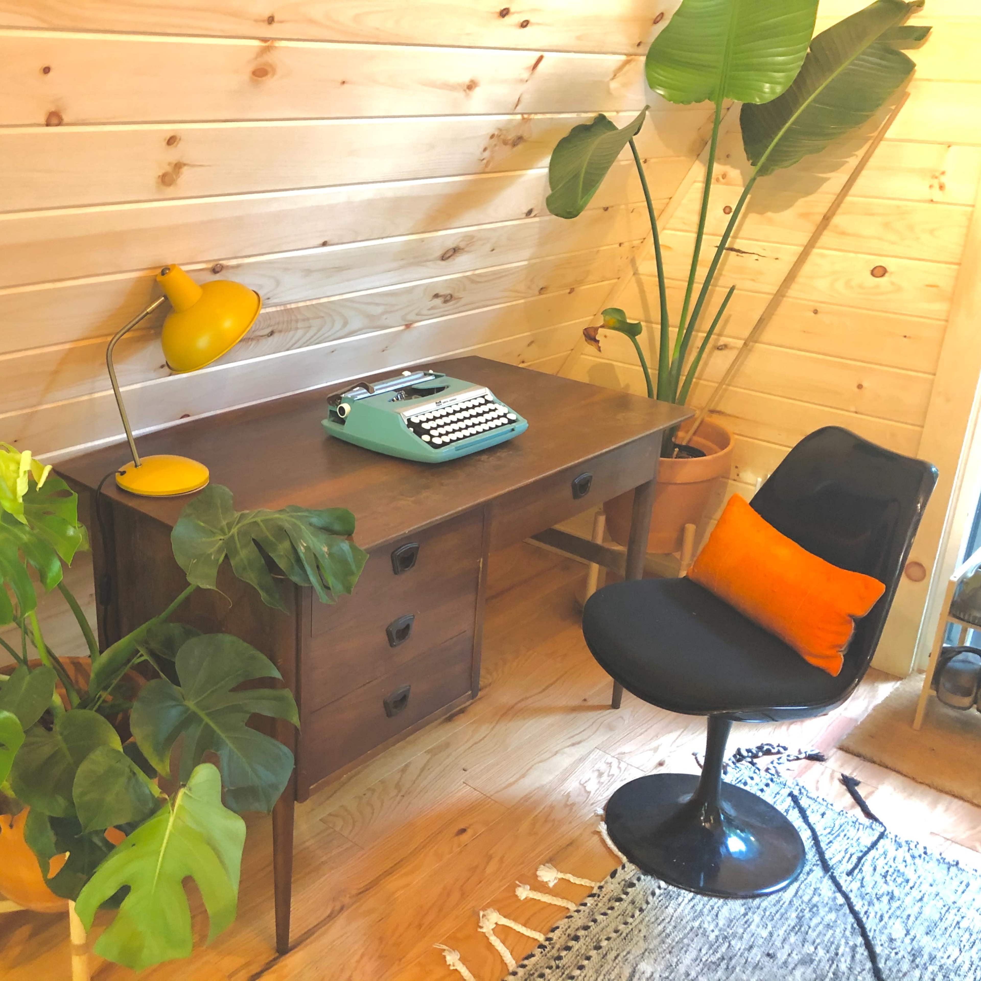 A wooden desk with a vintage typewriter is positioned next to a potted plant and a black chair in a room with wooden walls.