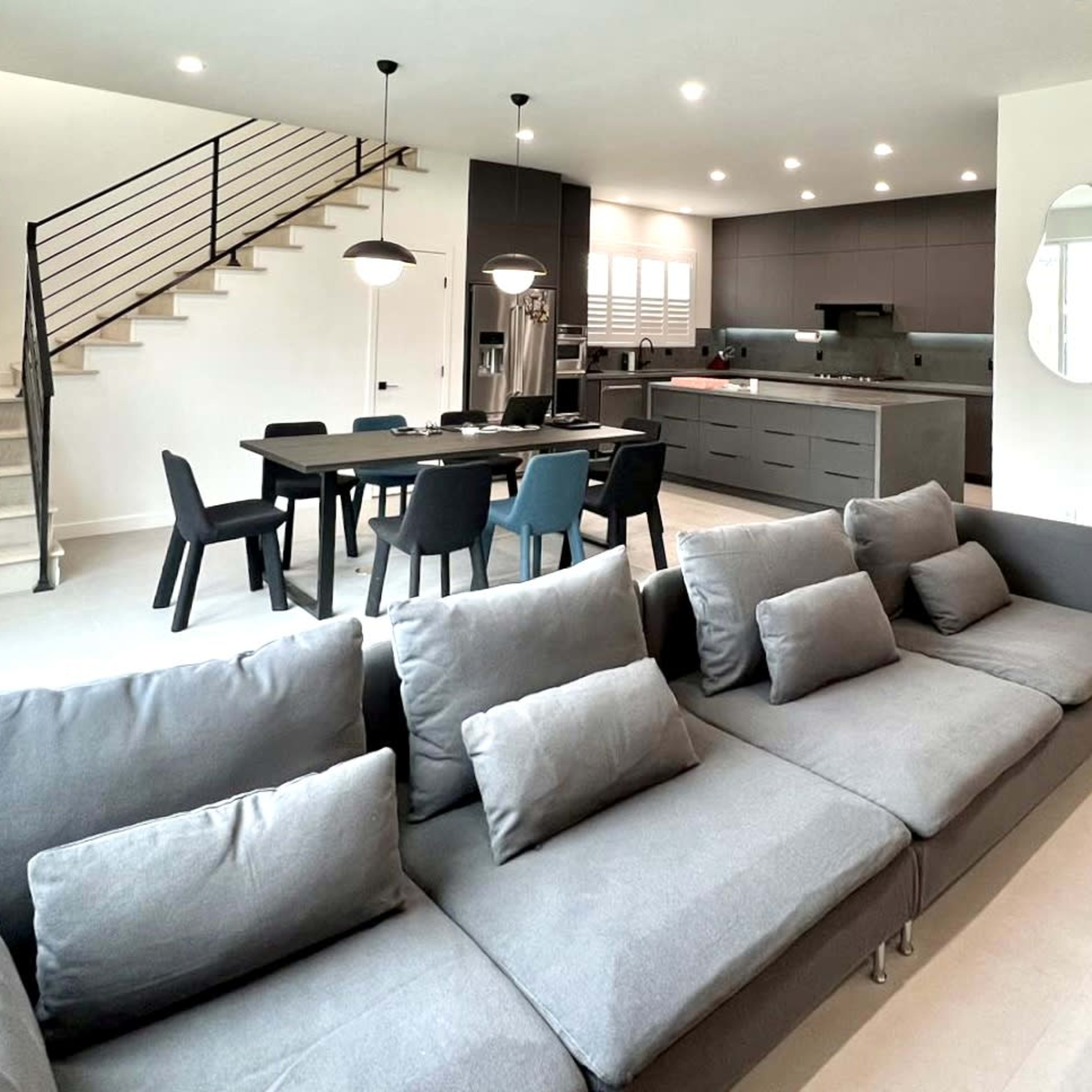 A modern living space with a grey sofa in the foreground, a dining area with a black table and chairs, and a kitchen featuring sleek cabinetry and appliances.