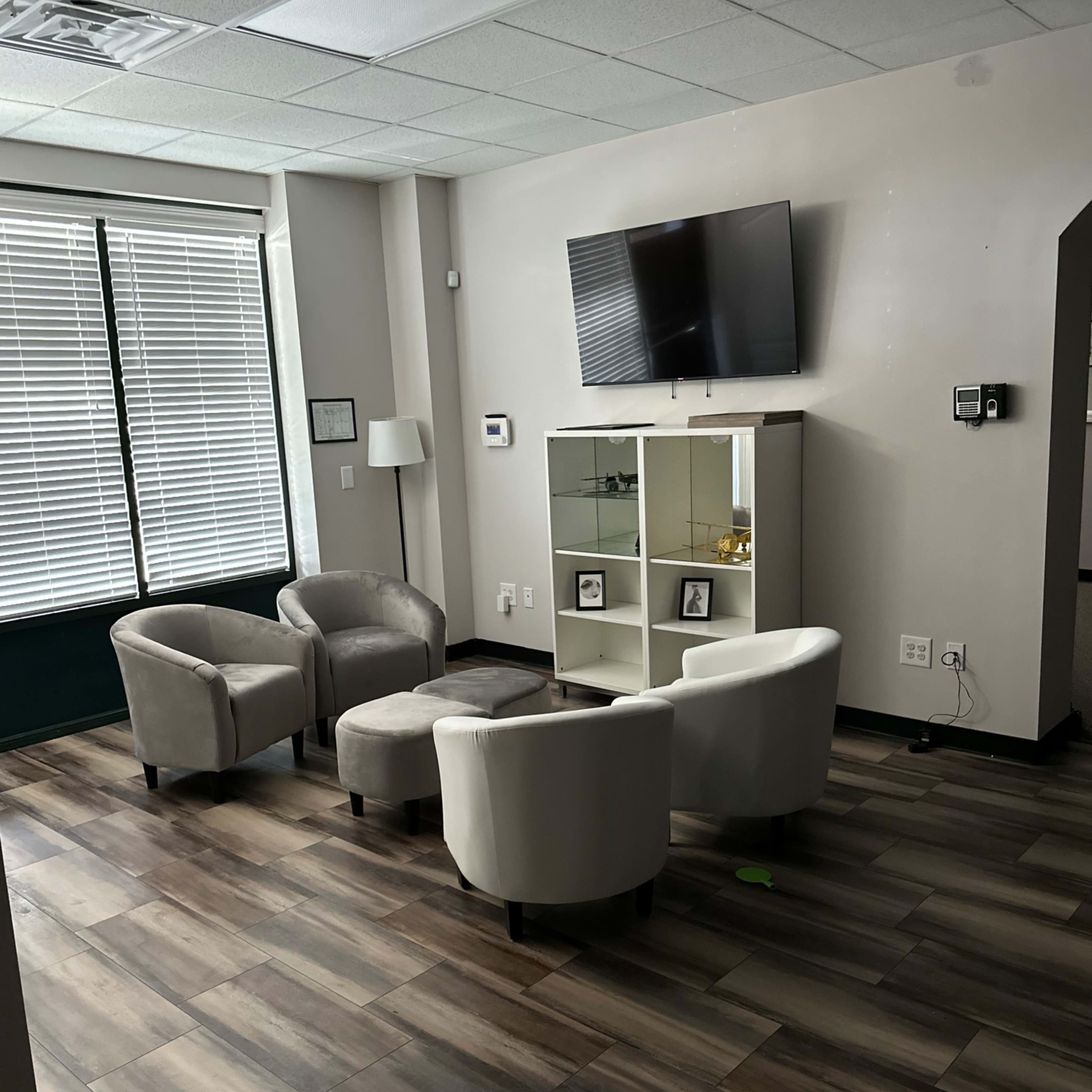 A waiting area with four light-colored chairs arranged around a small ottoman, a television mounted on the wall, and a glass cabinet displaying picture frames.