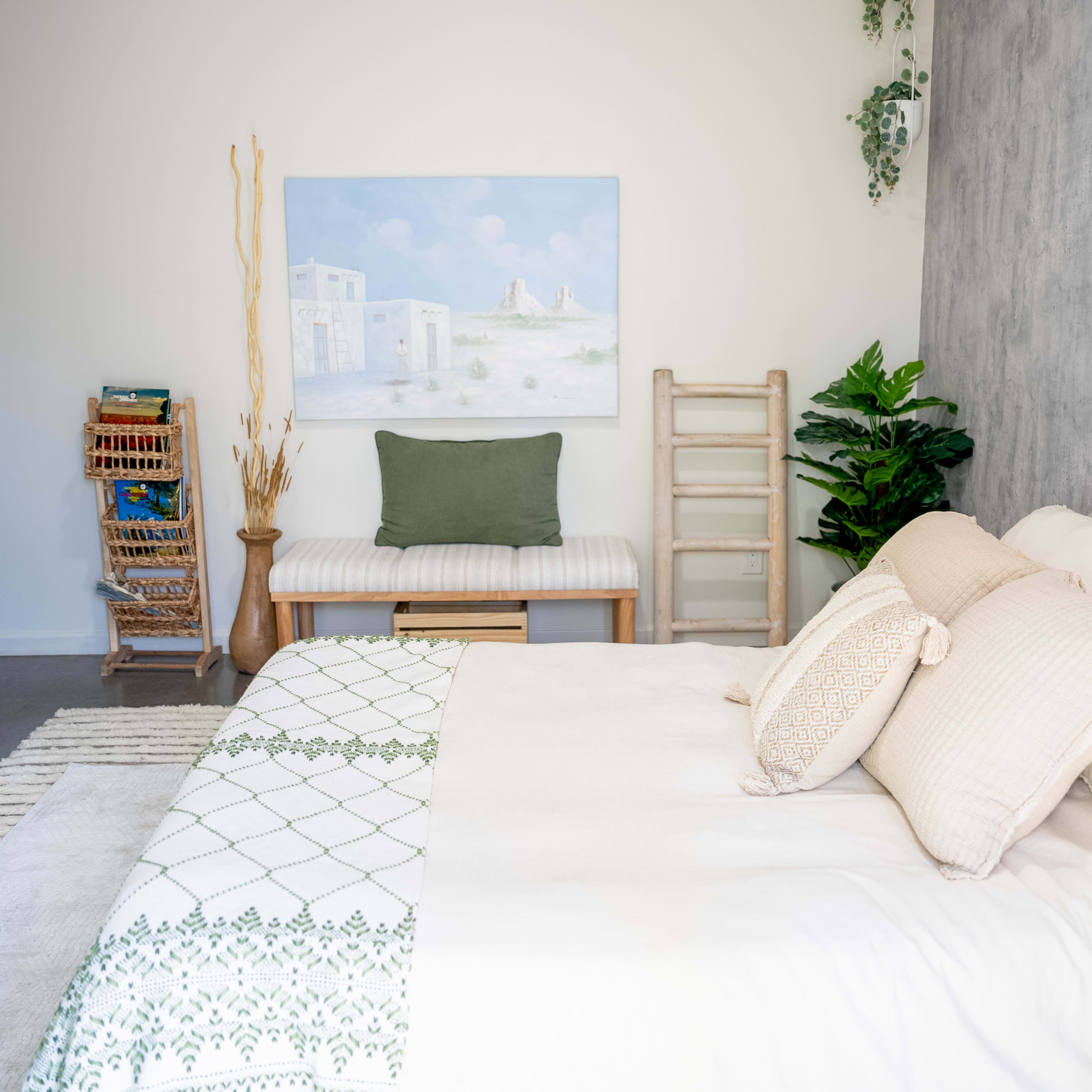 A well-organized bedroom features a large bed with light linens, a wooden ladder against the wall, and a painting above a bench.