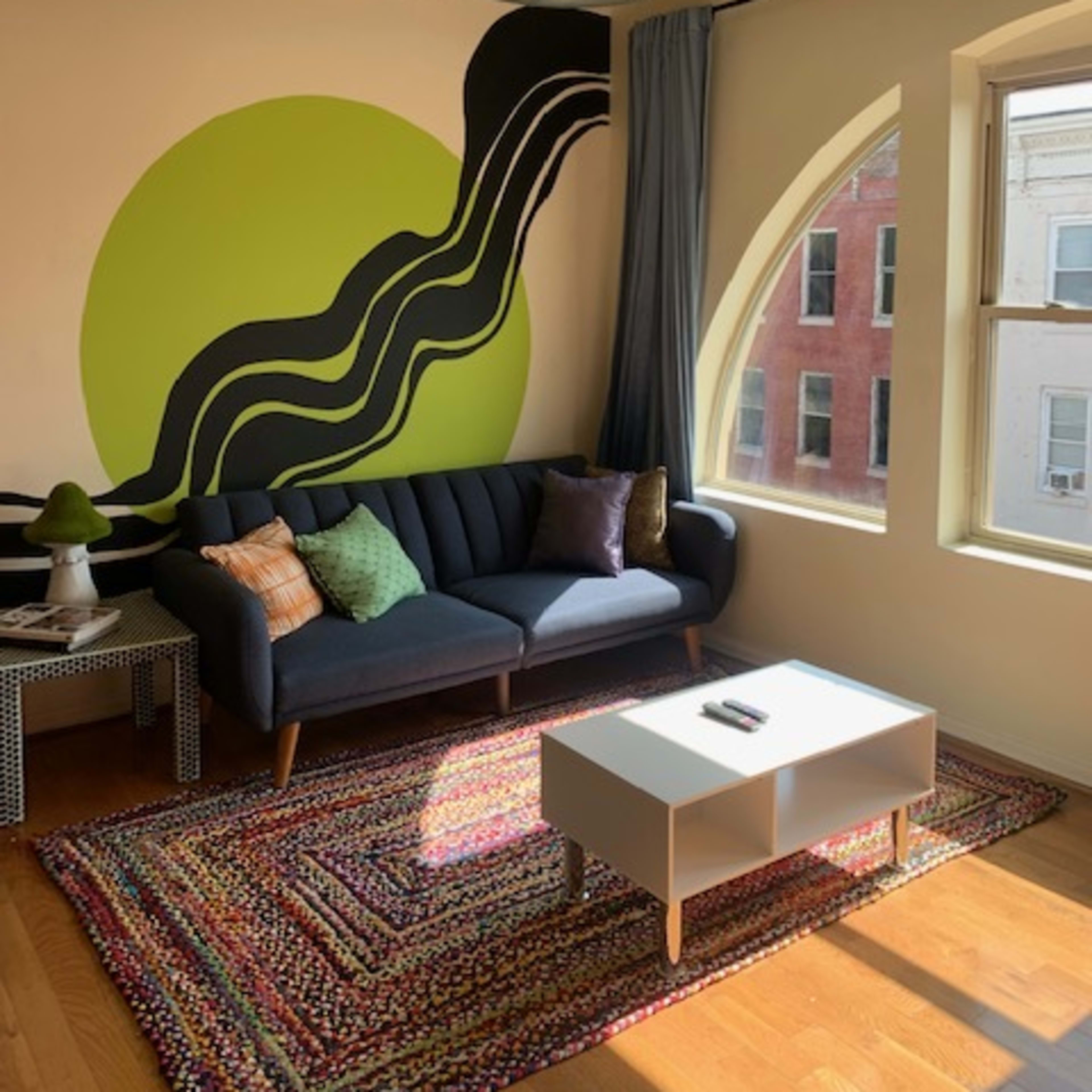 A modern living room with a dark sofa, a colorful area rug, and a large wall mural featuring bold green and black shapes.