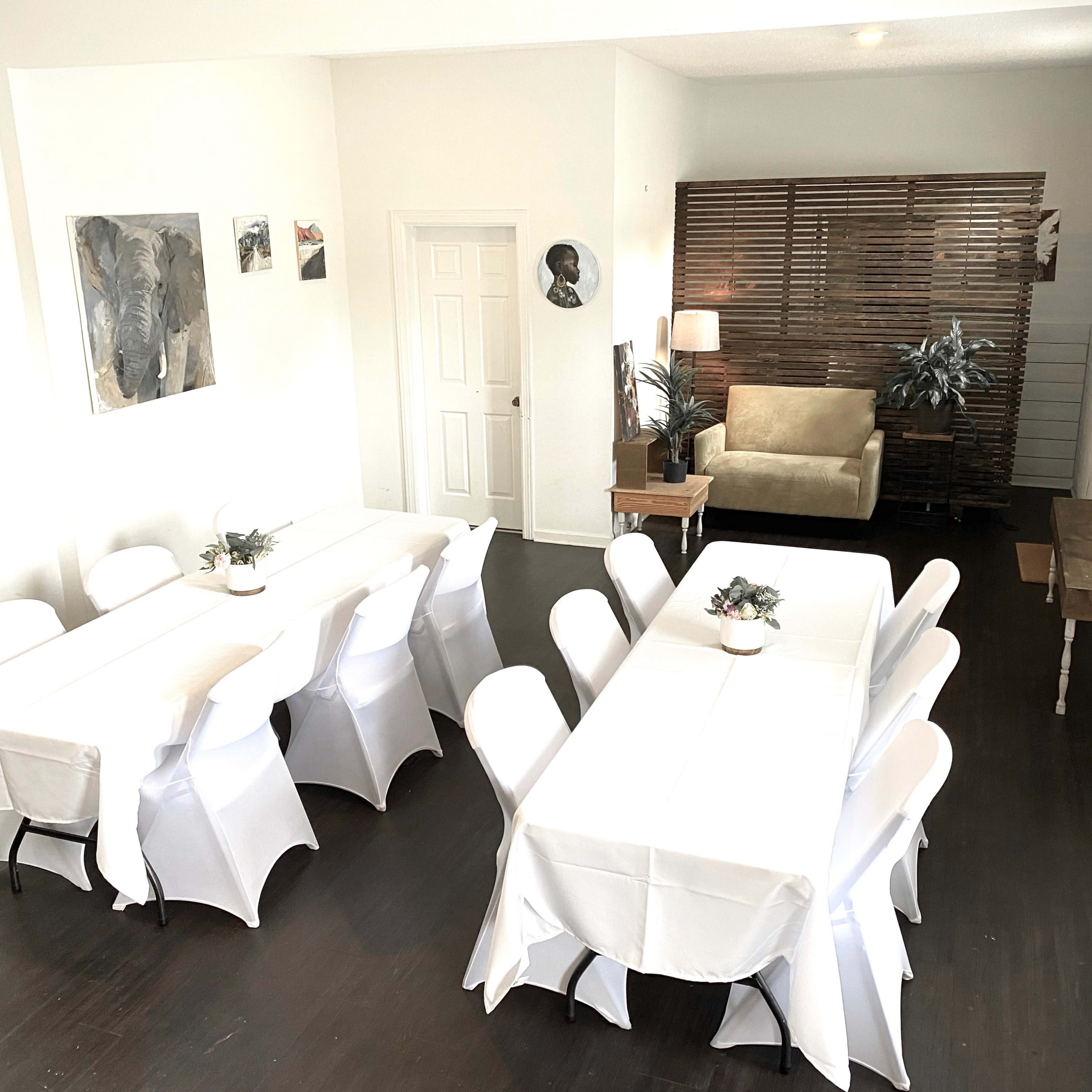 A spacious room features two long tables covered with white tablecloths, surrounded by white chairs, with a seating area and decorative artwork on the walls.