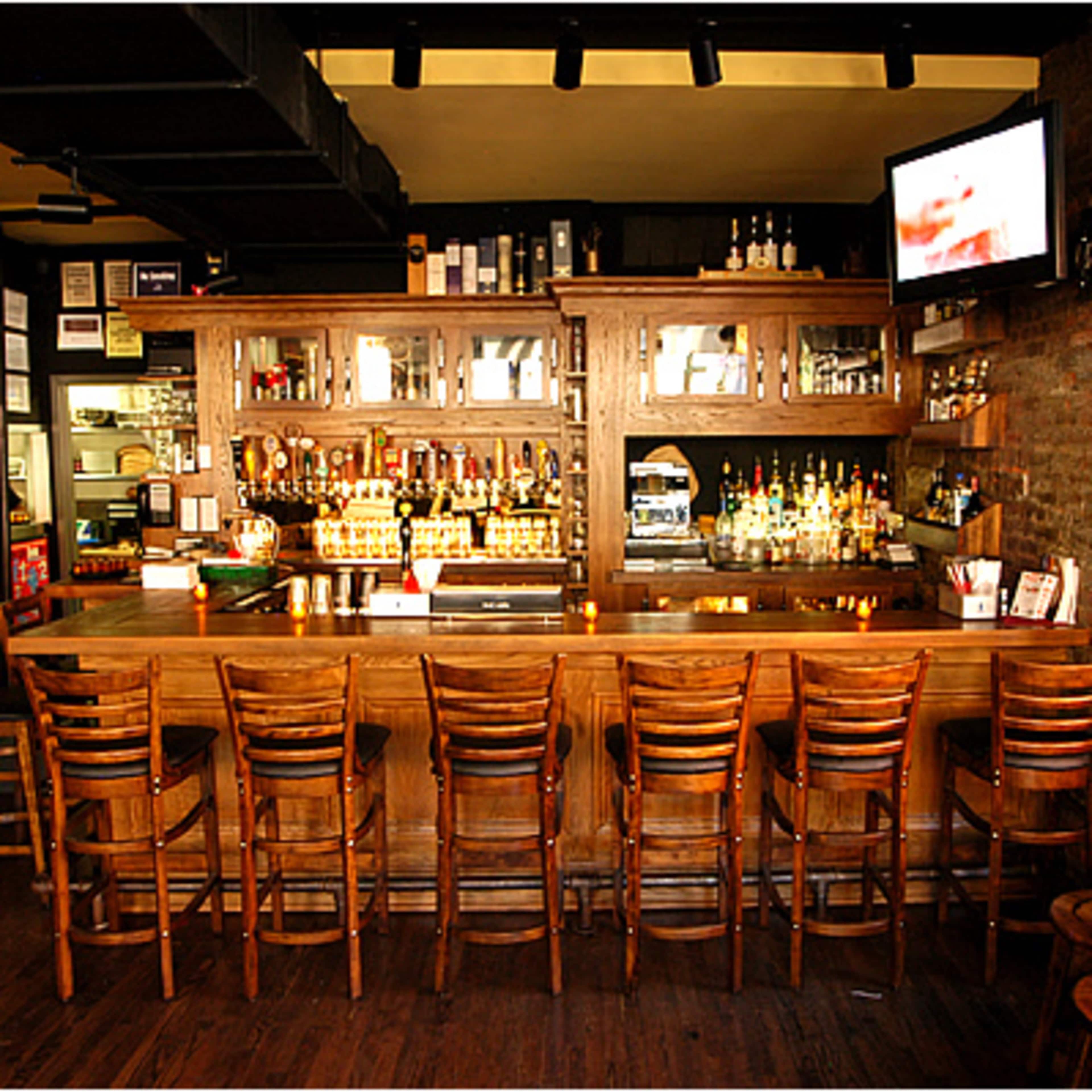 A bar with a wooden counter, several high stools, and shelves stocked with various bottles of liquor against a brick wall.