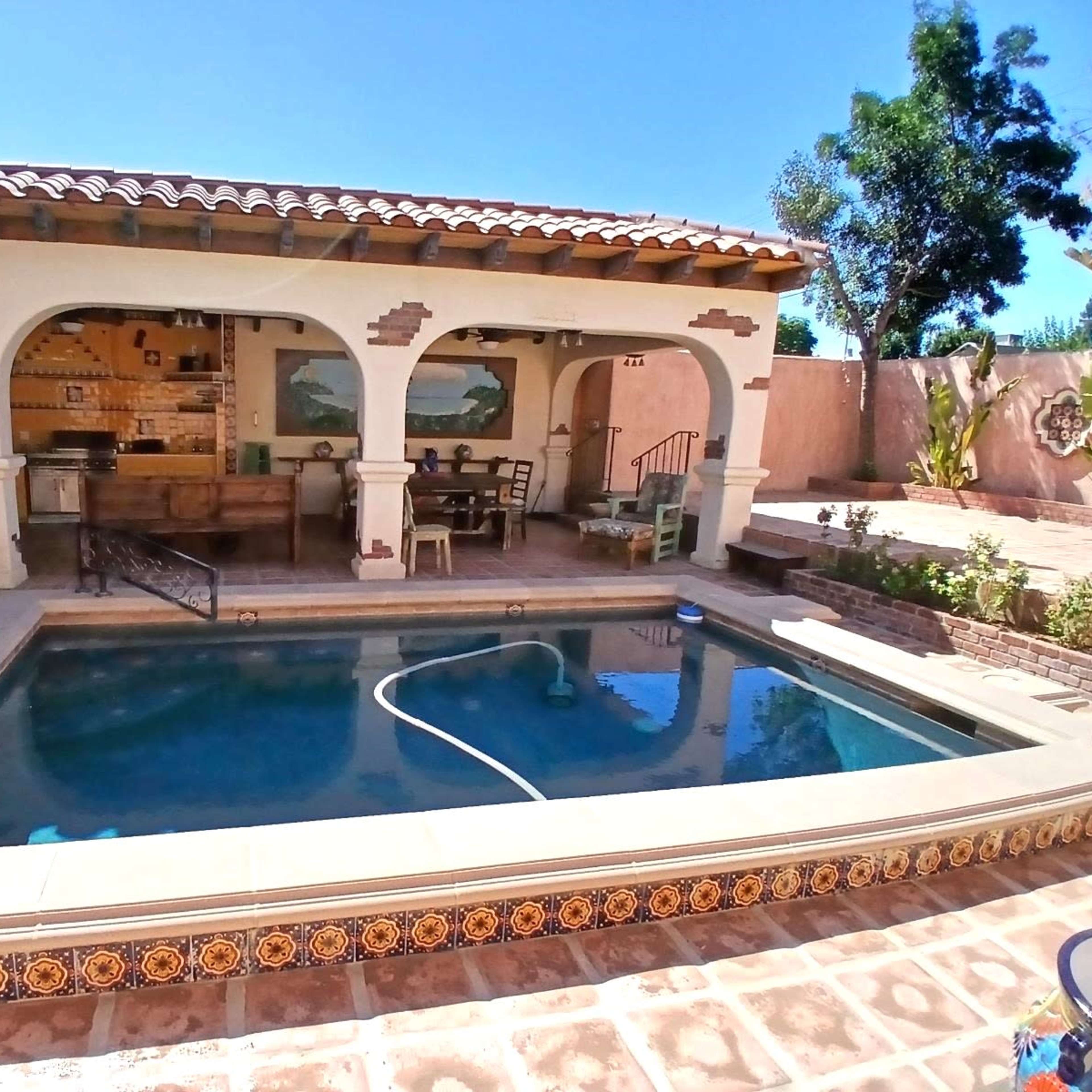 A courtyard with a pool, surrounded by a tiled patio and a covered outdoor seating area that includes a kitchen and dining space.