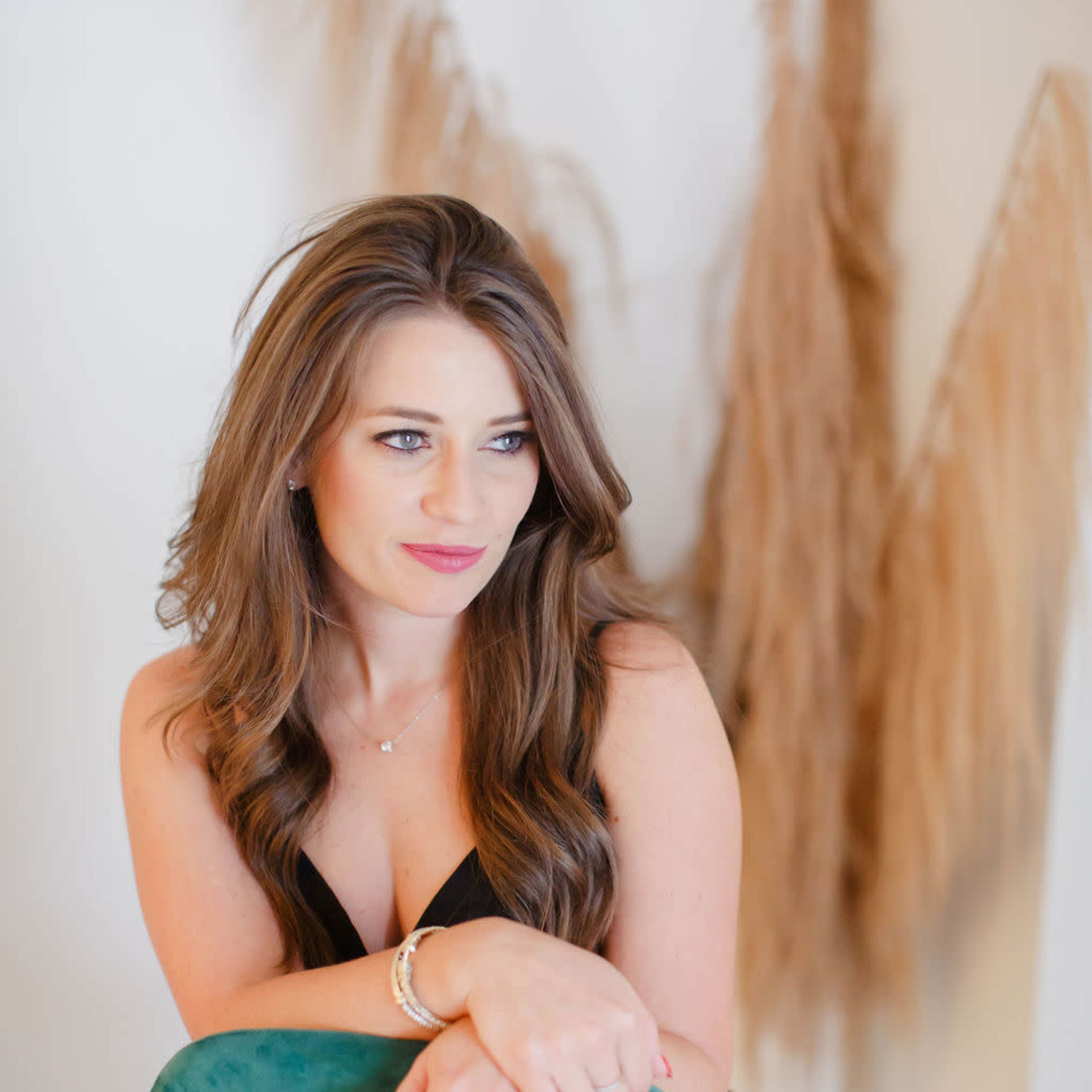 A woman with long hair sits elegantly on a teal chair in front of a pale wall adorned with dried decorative grasses.